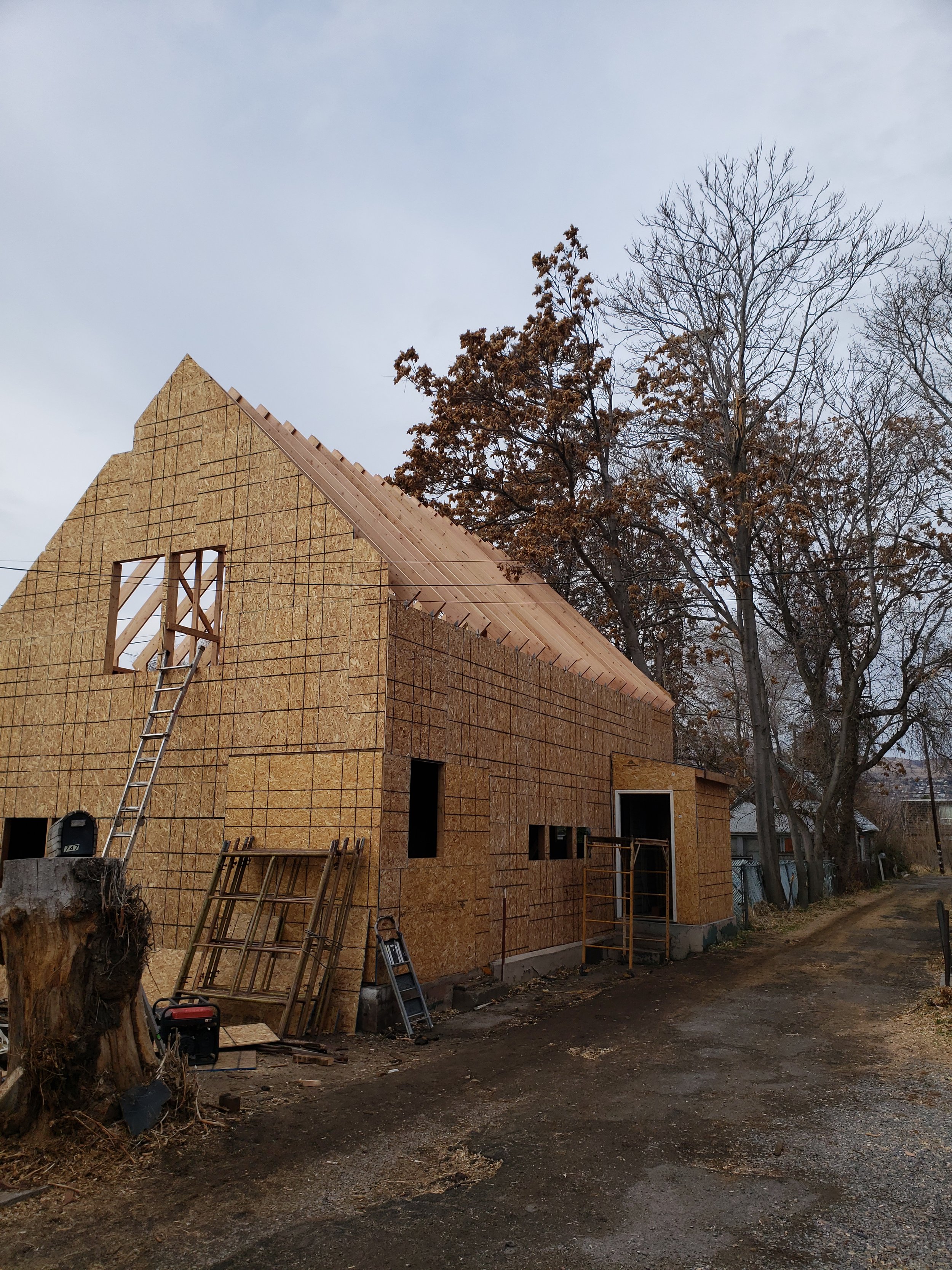 Construction of a house with wooden framing and plywood sheathing, on a dirt road with trees in the background.