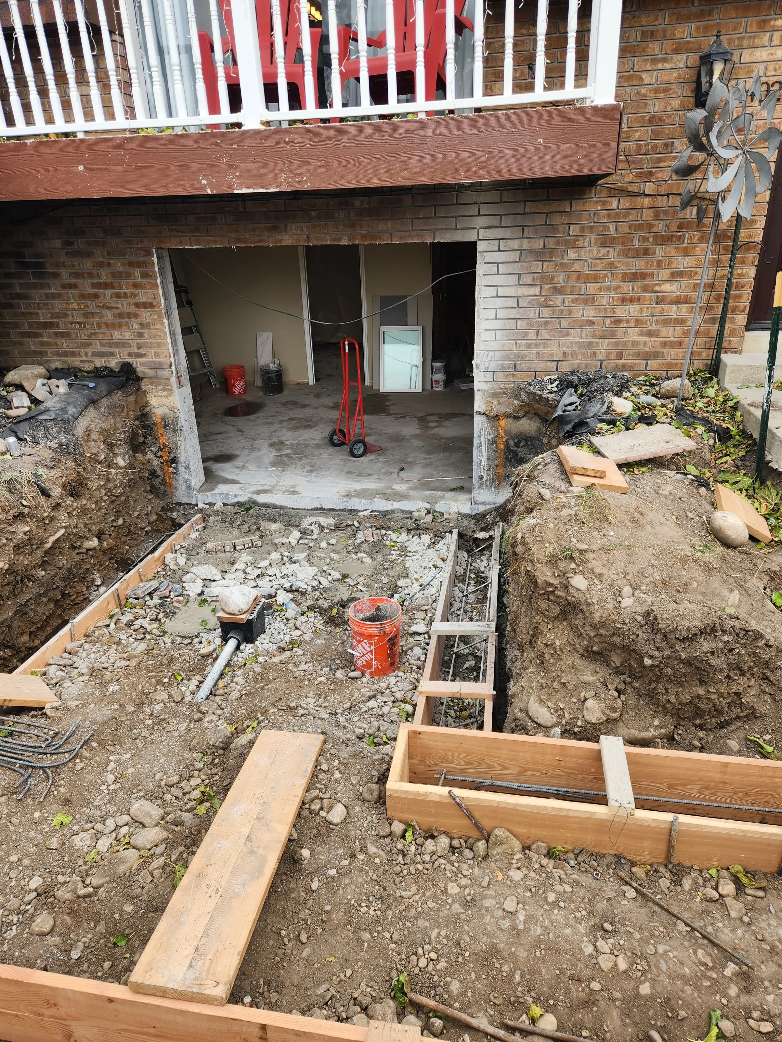 Construction site of an excavation in a backyard with exposed soil, wooden framing, construction tools, and building materials. The garage door opening is visible, leading to an indoor space with painting supplies and a ladder.