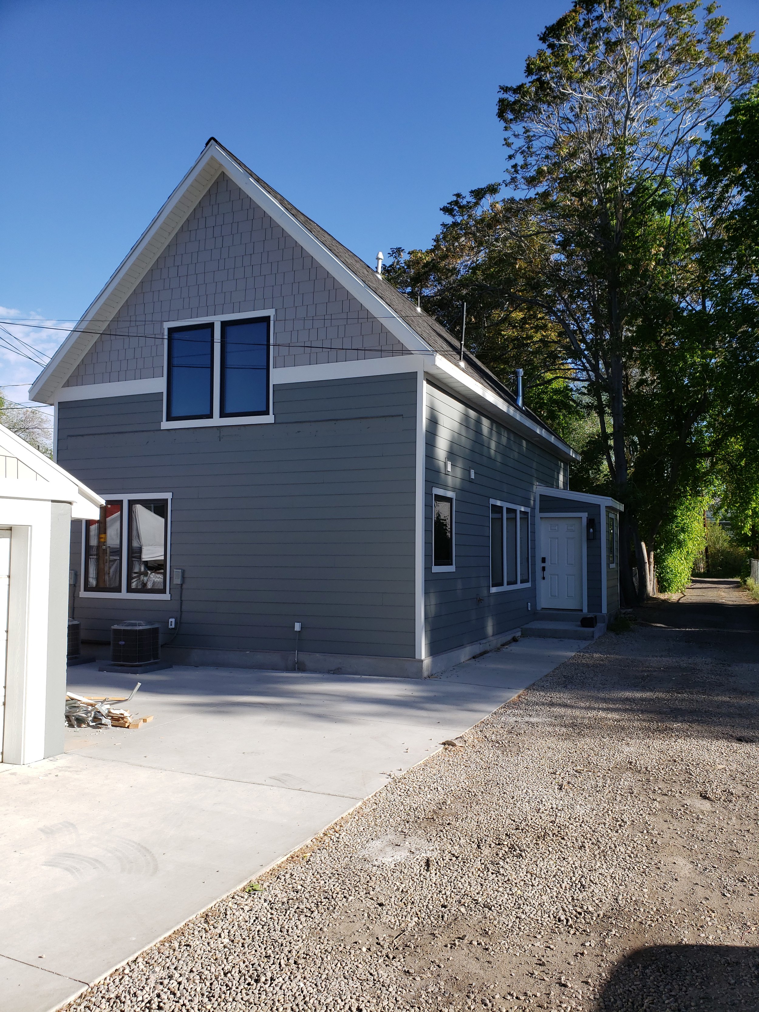 A new light gray house with white trim, two stories, large windows, and a gable roof. A small concrete step leads to the front door, and there's a concrete driveway next to the house. Trees with green leaves are in the background under a clear blue s