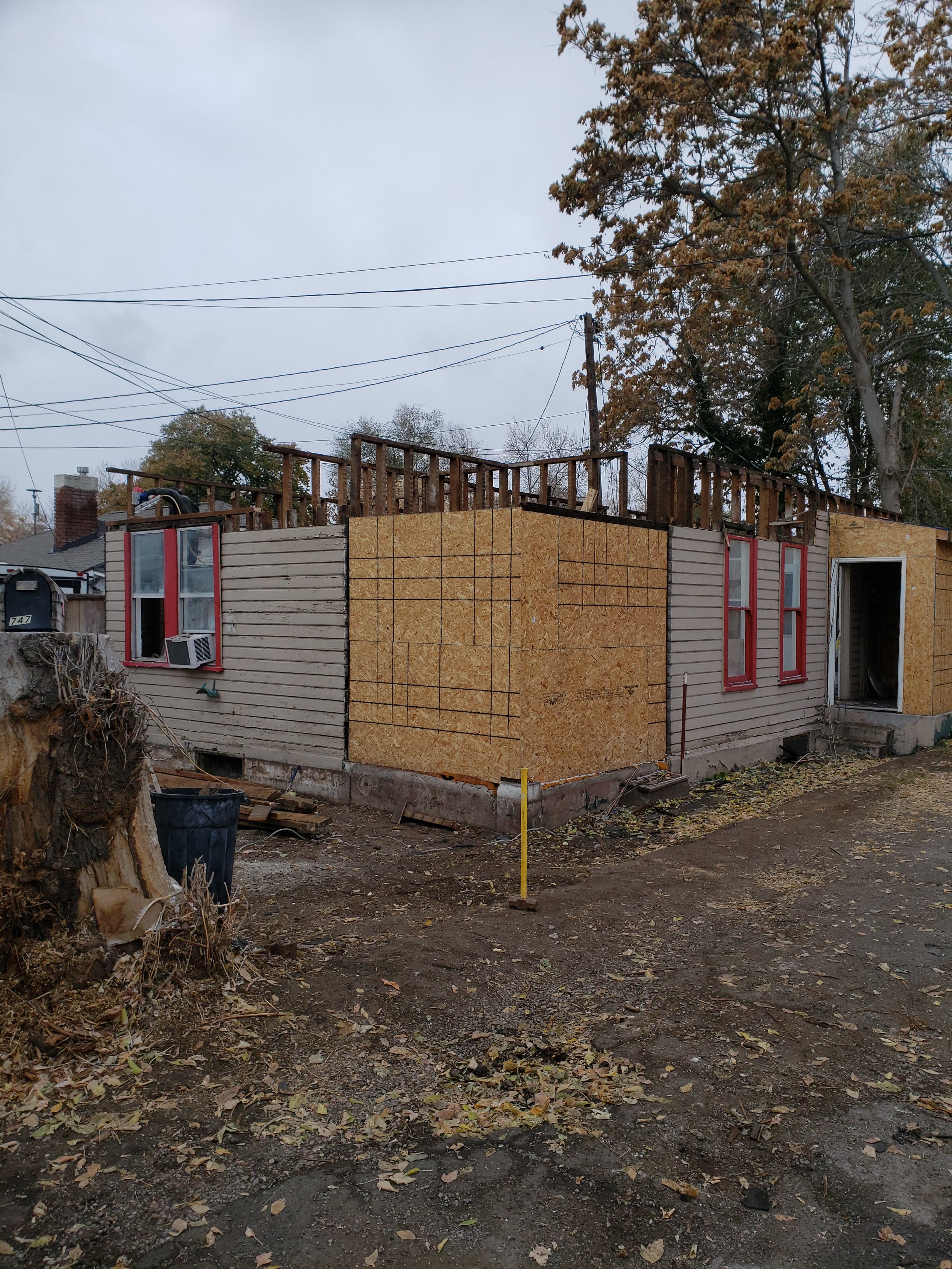 House under construction with boarded-up exterior, partially built upper floor, and trees in the background.