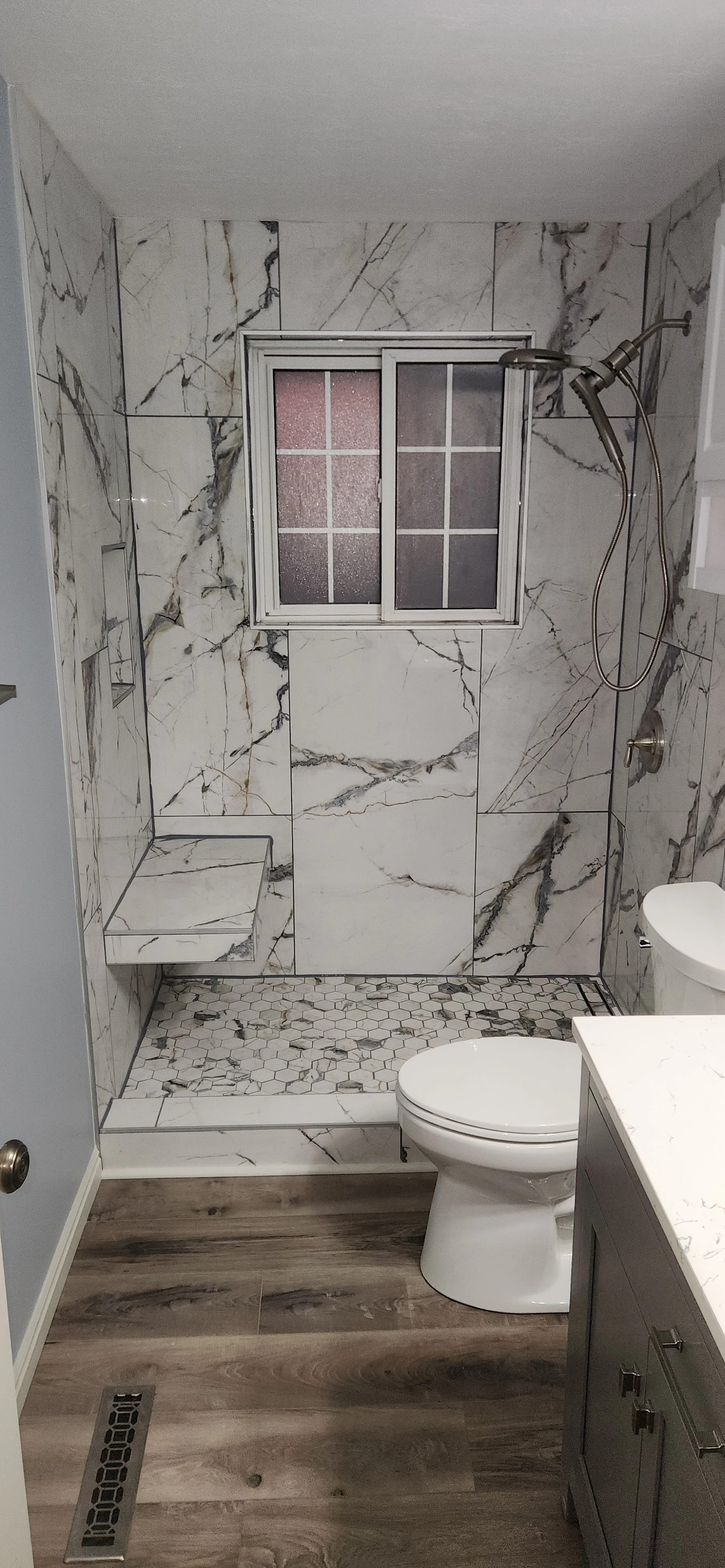 Bathroom with a walk-in shower featuring marble tiles, a window, a toilet, and a vanity with a marble countertop. The shower has a built-in bench and a handheld showerhead, with a hexagon and marble pattern on the floor.