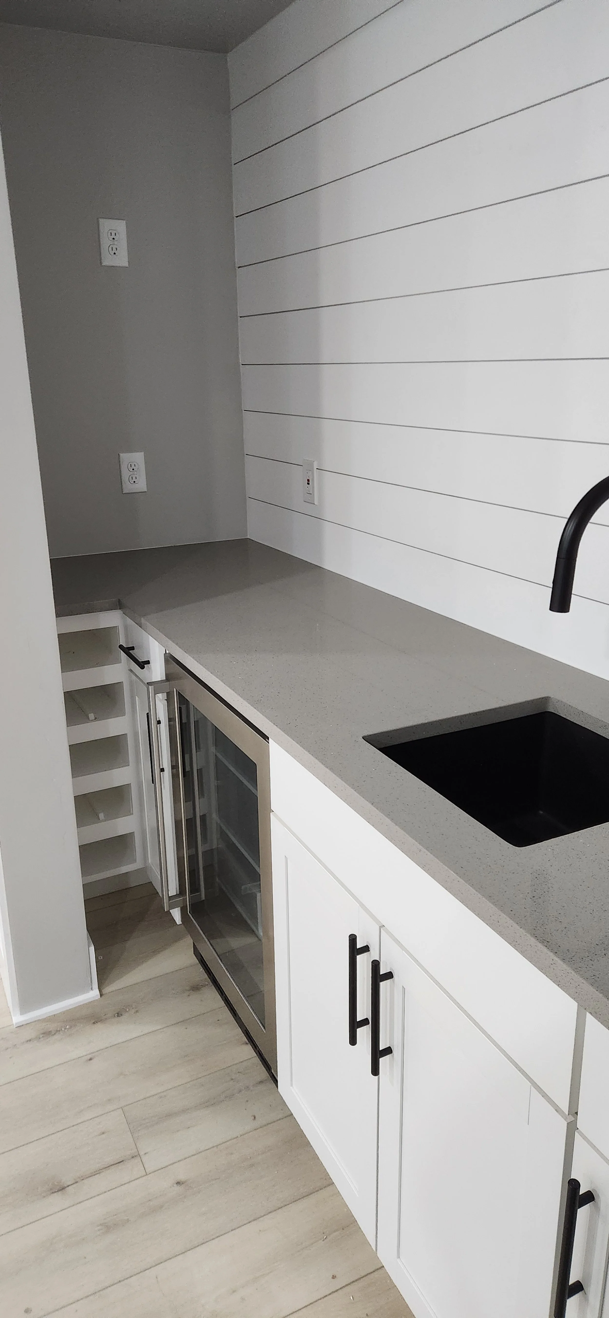 Kitchen area with white cabinets, black faucet, black sink, mini wine fridge, and electrical outlets, with wood-like flooring and a gray countertop.