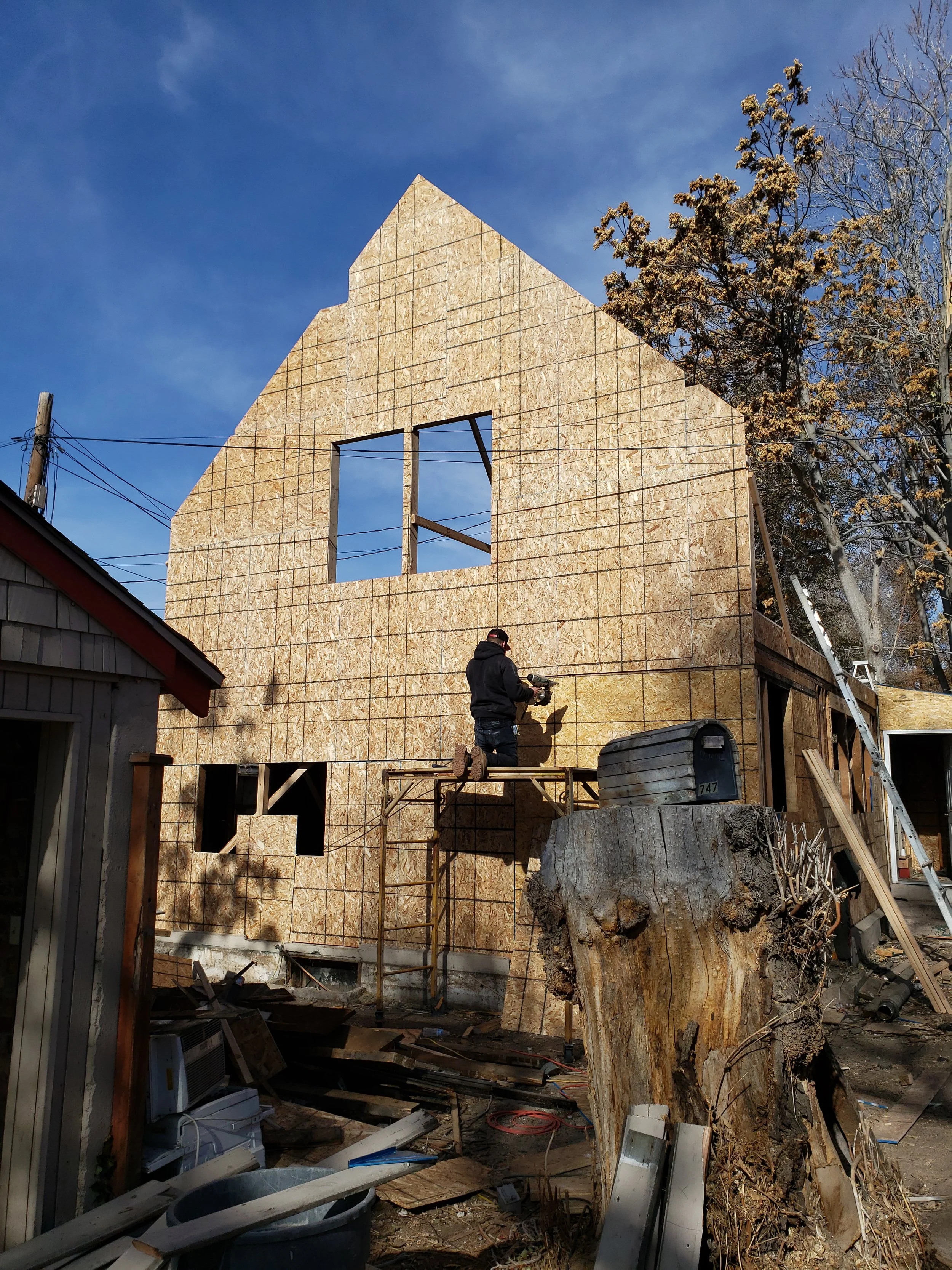 Construction worker installing plywood wall on a house under construction, with trees and blue sky in the background.