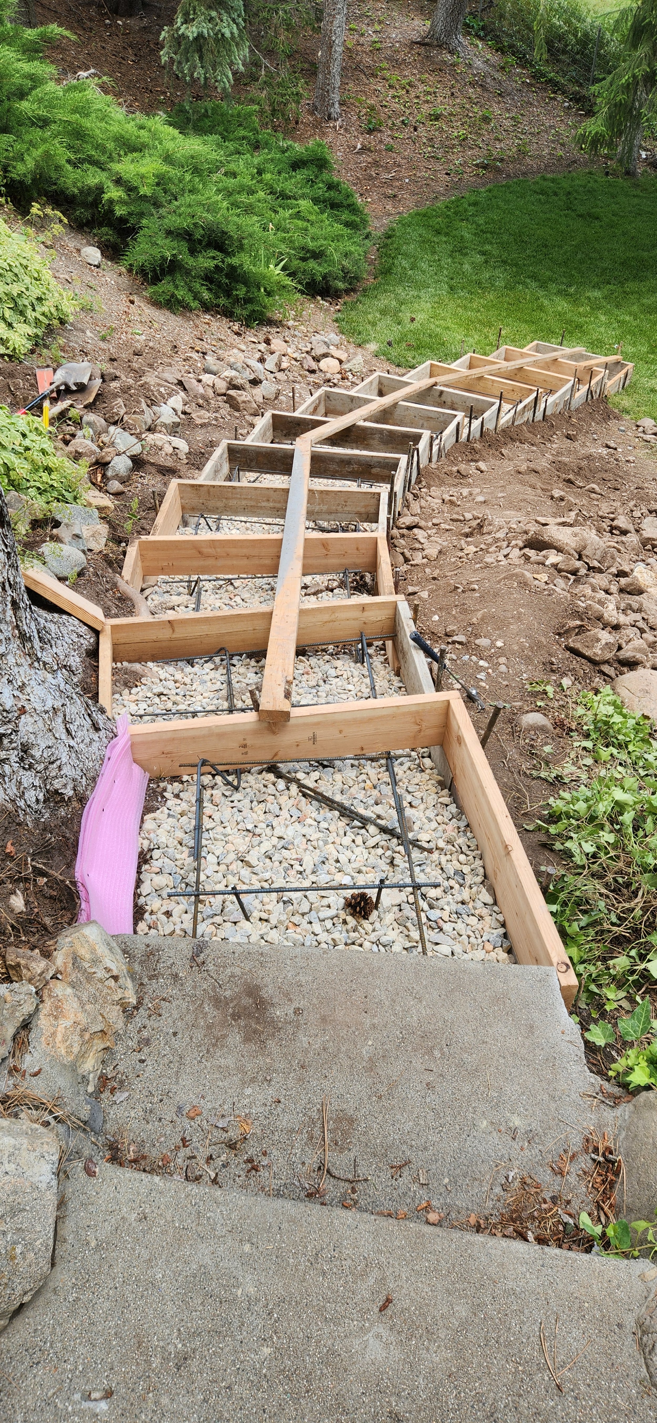Wooden formwork and rebar for a garden or pathway trim being constructed on a sloped yard, with a mix of grass, plants, and trees surrounding the area.