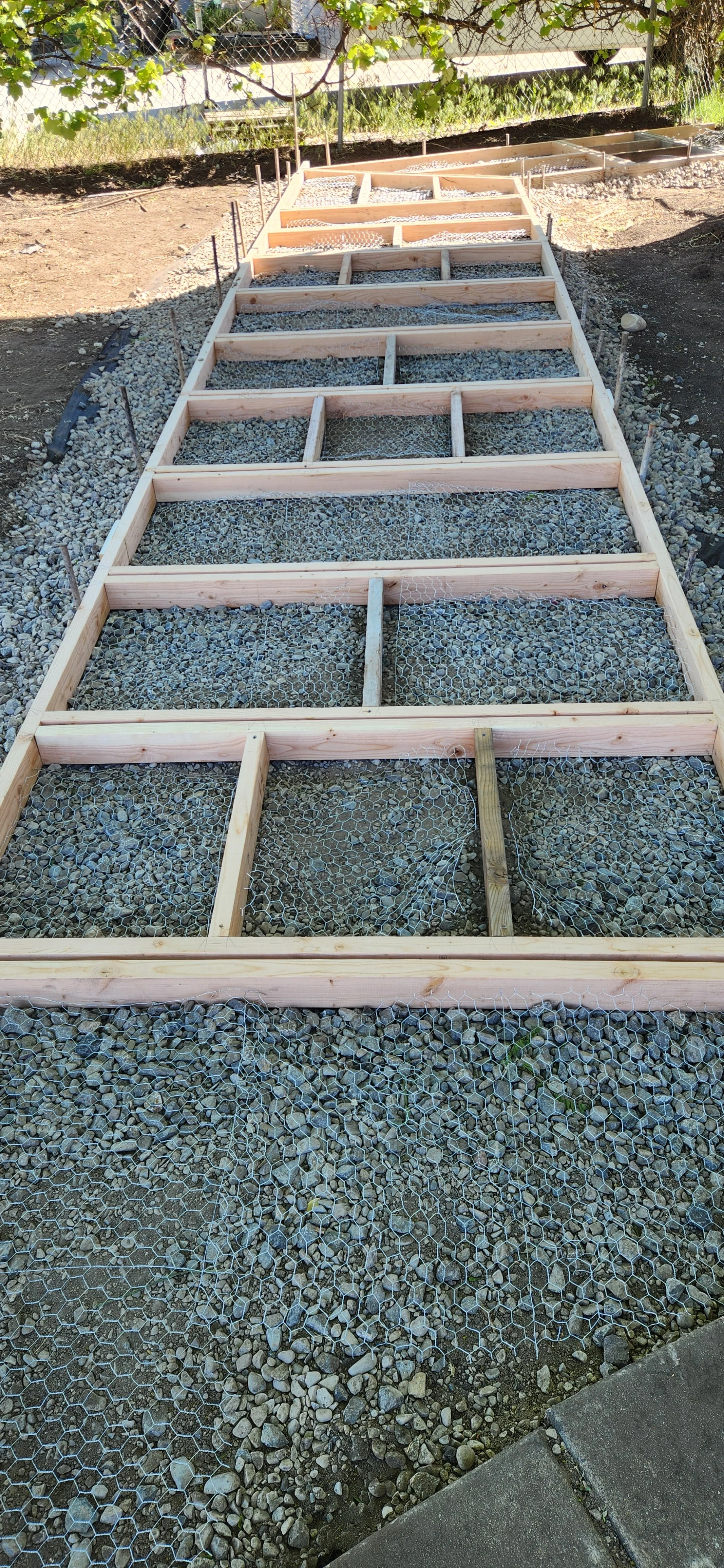 Wooden framing structure for a garden pathway or raised bed being built on gravel, with chicken wire attached.