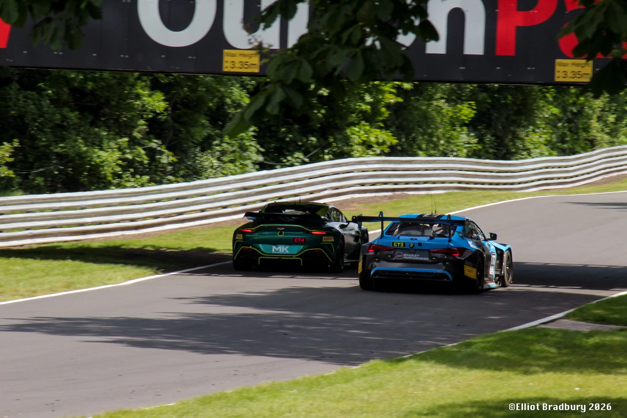 Two race cars, one black and green with 'MK' and 'GT4' markings, and one blue with 'GT3' markings, on a racetrack surrounded by greenery and a metal guardrail, during a race.
