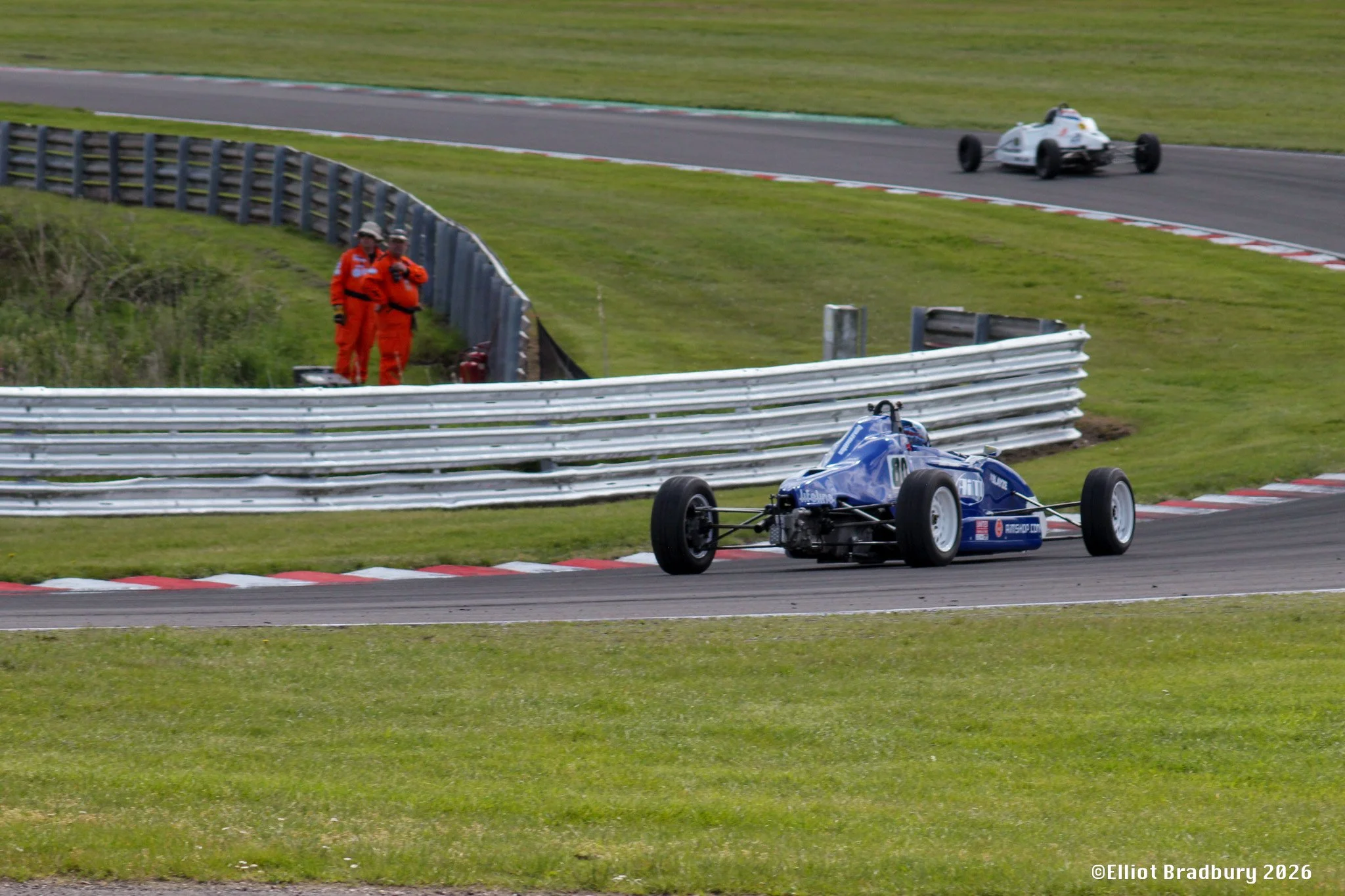 Racecar on a race track with two marshals in orange suits watching from the side.