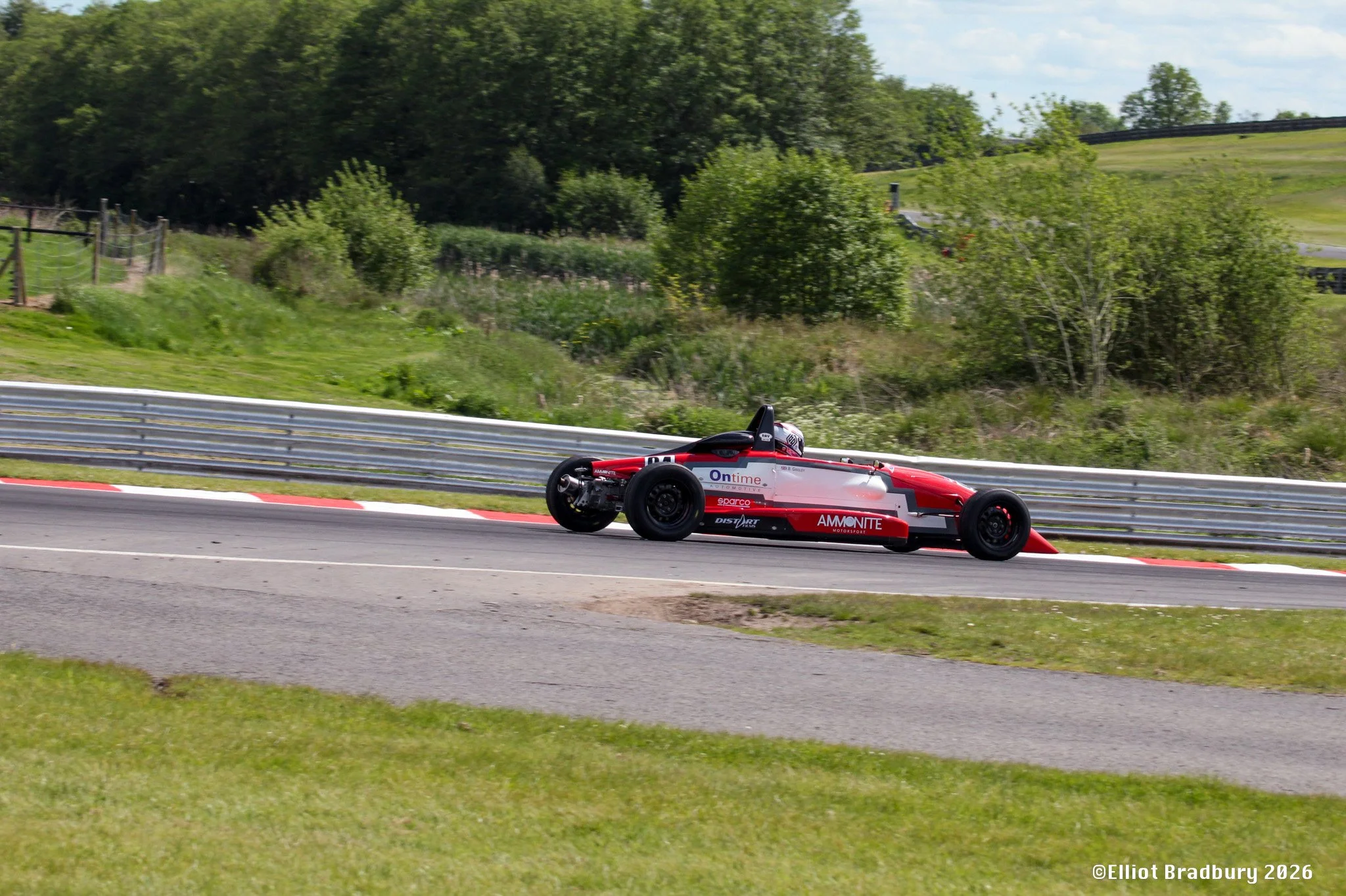 Red and white racing car on a race track, with green trees and grassy hills in the background.