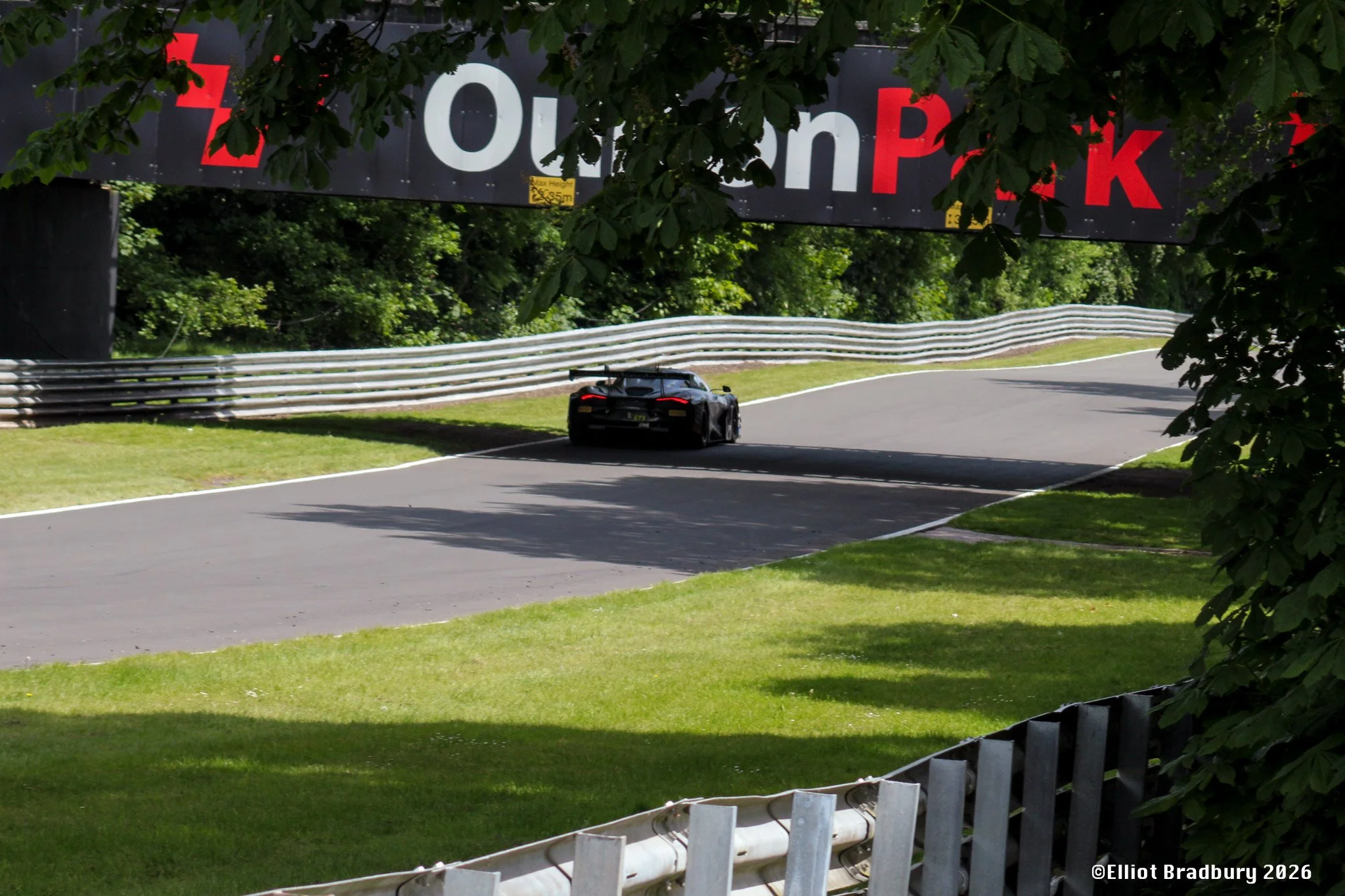 A black race car driving on a winding road surrounded by trees and greenery, with a large black and red sign overhead that reads 'Oulton Park'.