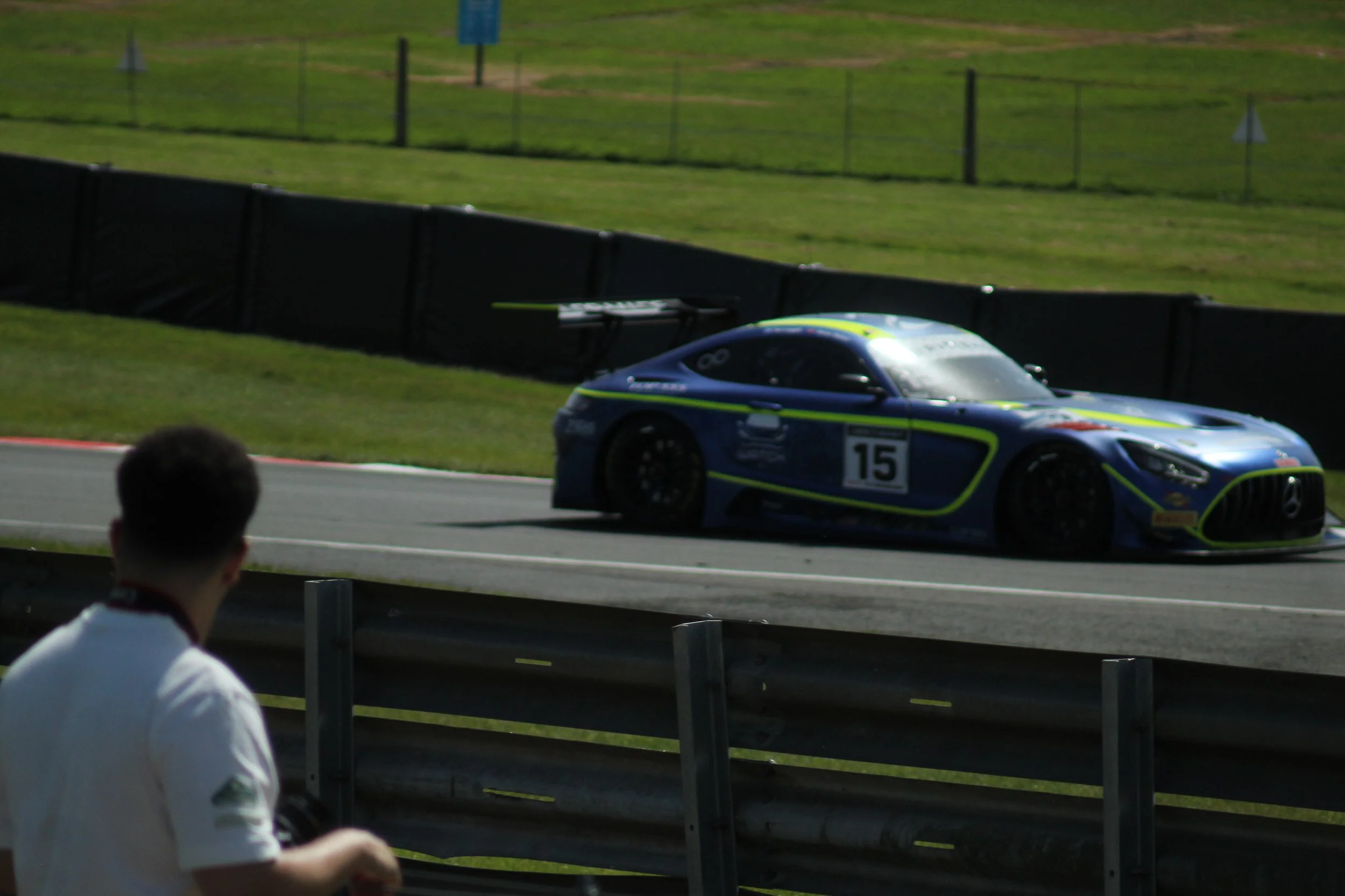A blue race car with the number 15 on its side speeds along a racetrack, with spectators watching from behind a barrier, and a grassy area with some signs in the background.