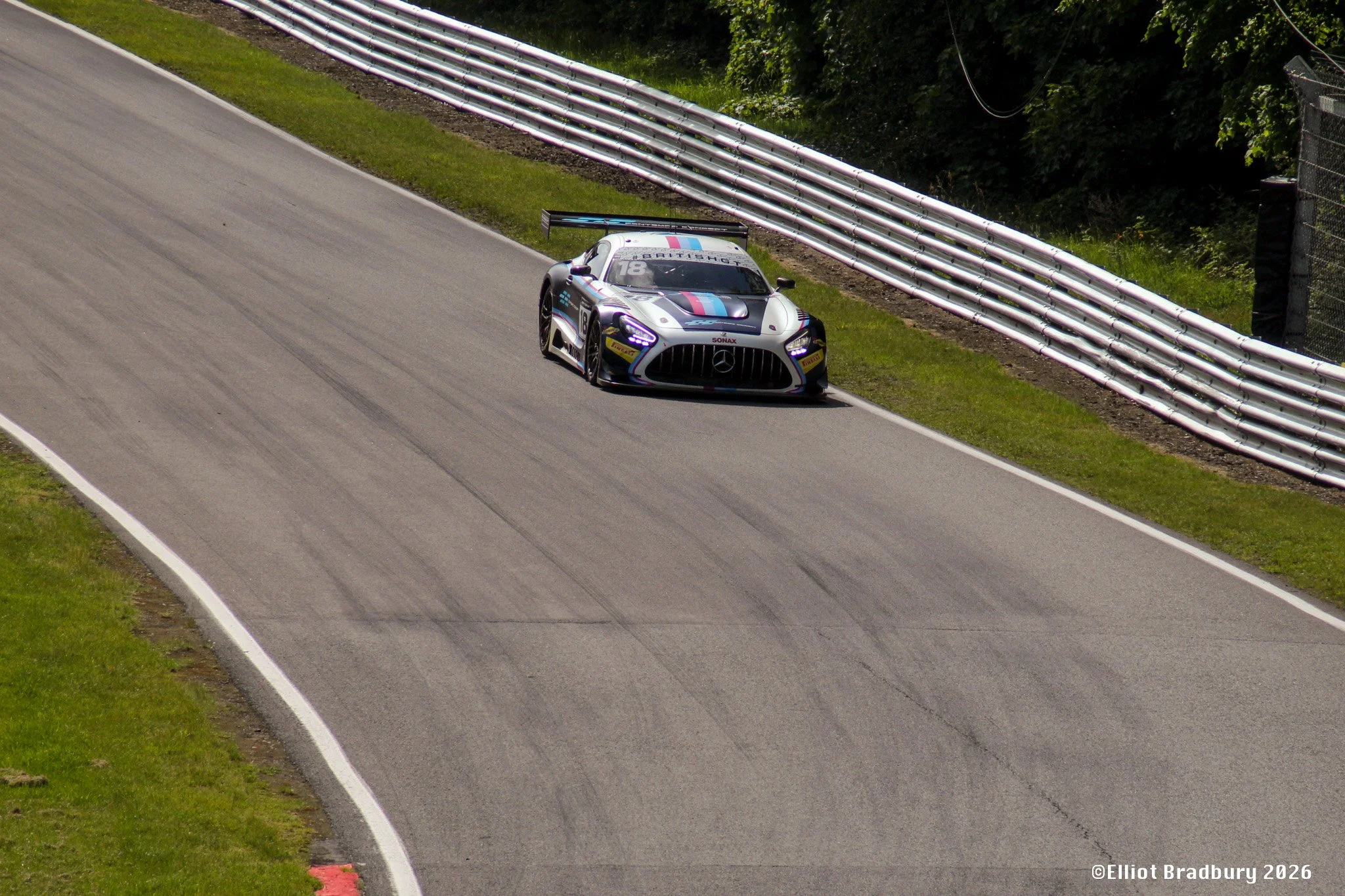A race car on a winding gravel race track, with metal barriers lining the edge and green trees in the background.