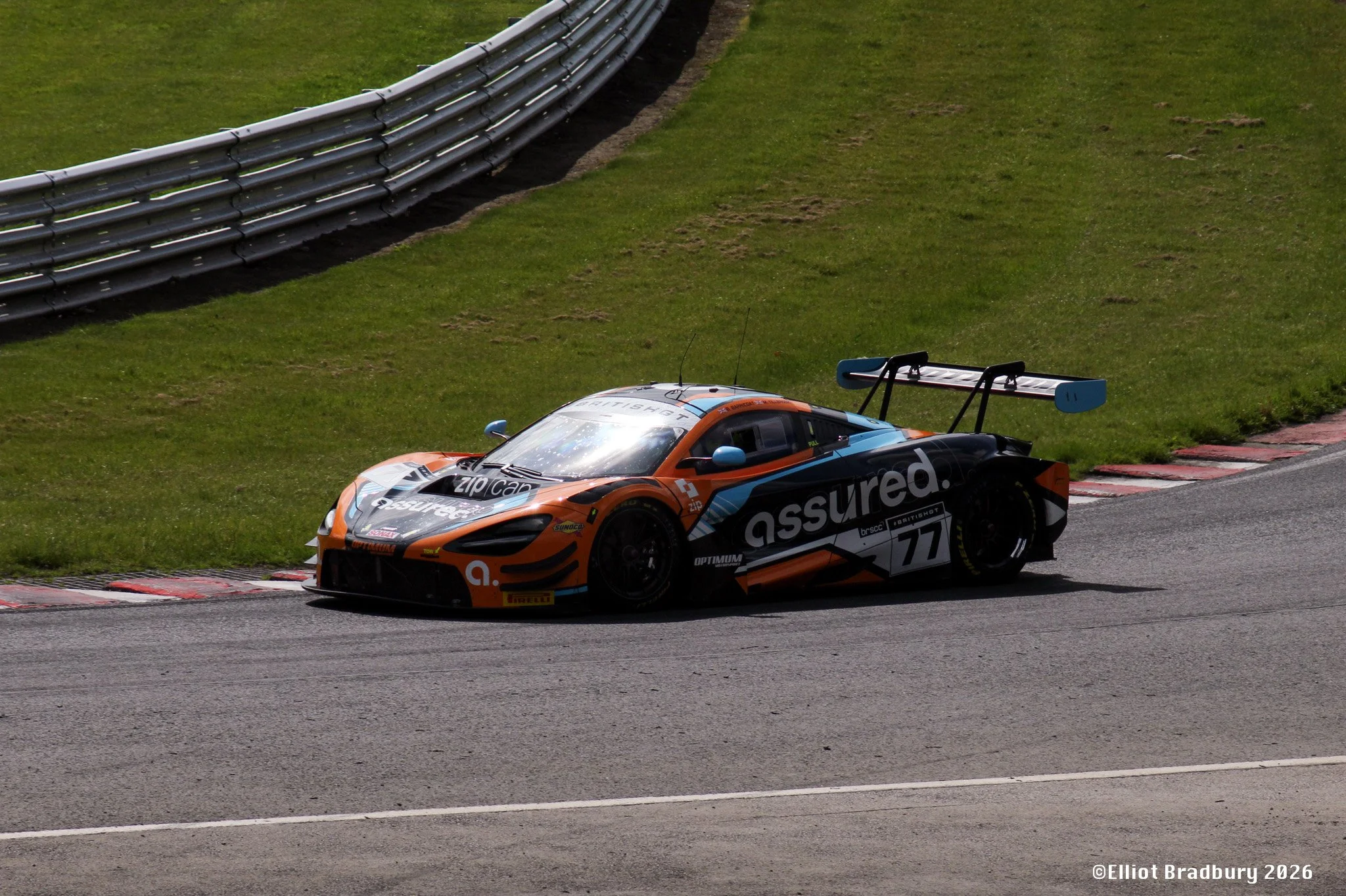 A race car on a racetrack, approaching a corner, with grass and tire barriers in the background.