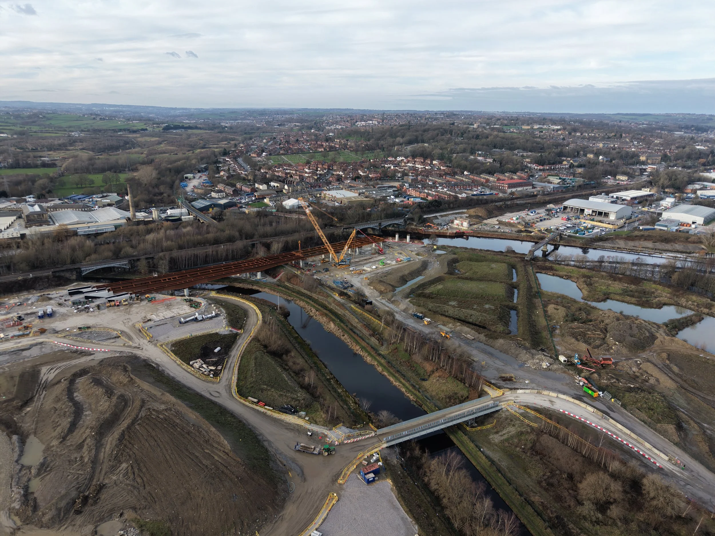 An aerial view of a construction site with cranes, roads, and water bodies, with a cityscape and farmland in the background.
