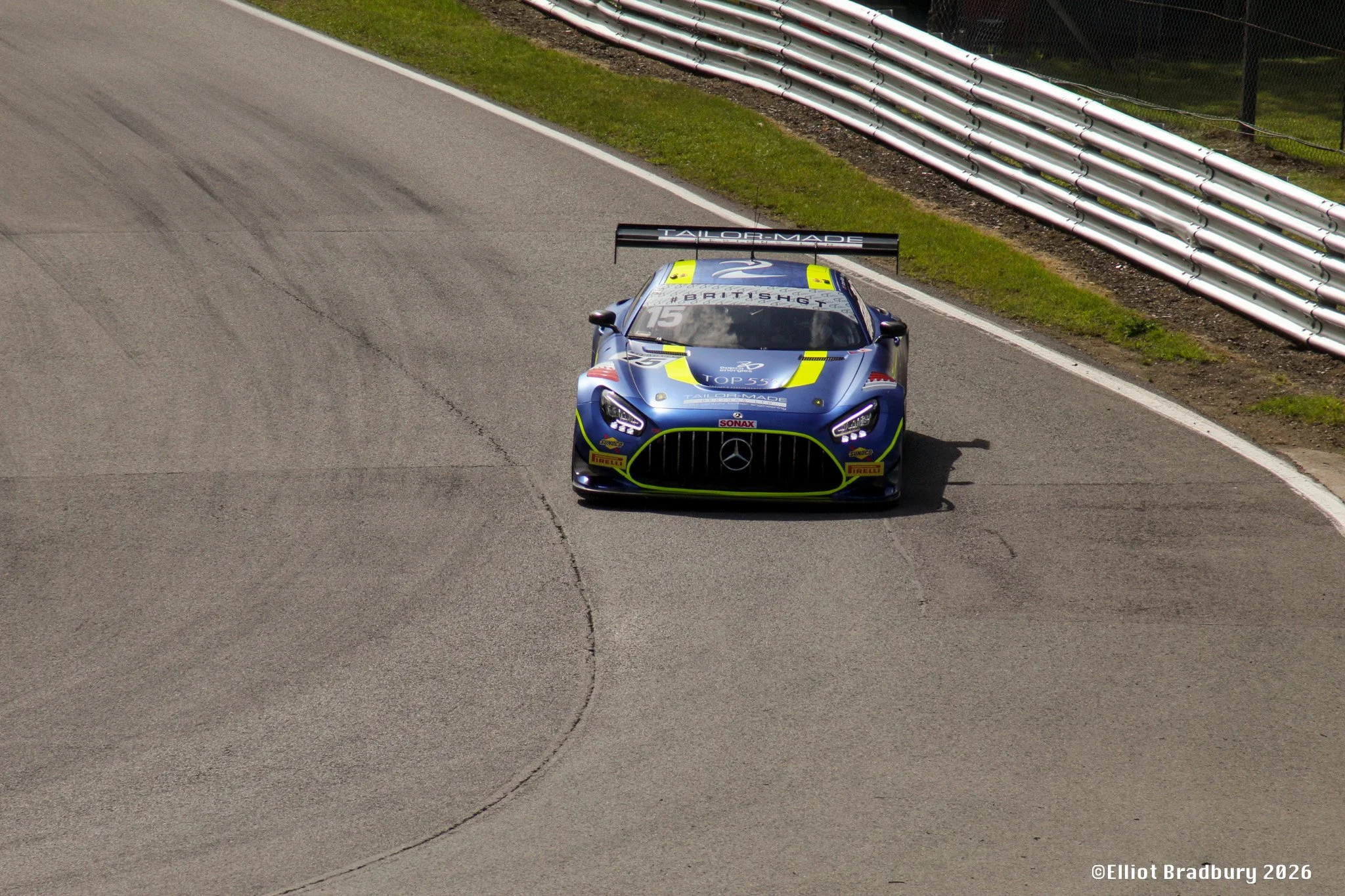 A blue mercedes race car on a racing track with a large rear wing that has the word 'TAILOR-MADE' on it, and various sponsor logos including Pirelli and Sonnax. The track has tire marks and is bordered by a grass verge and safety barriers.
