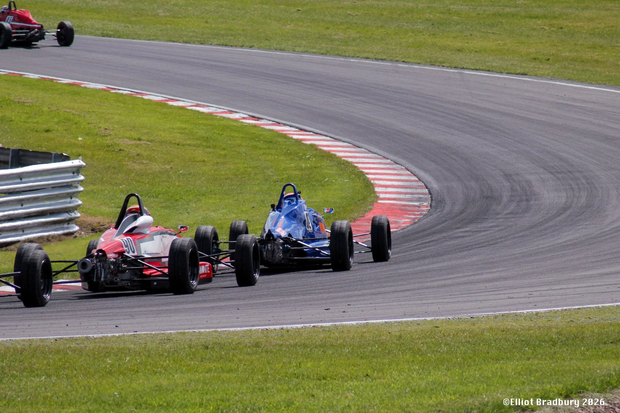 Two open-wheel race cars on a paved race track, with a red and white striped curb on the inside of the turn and grassy areas nearby.