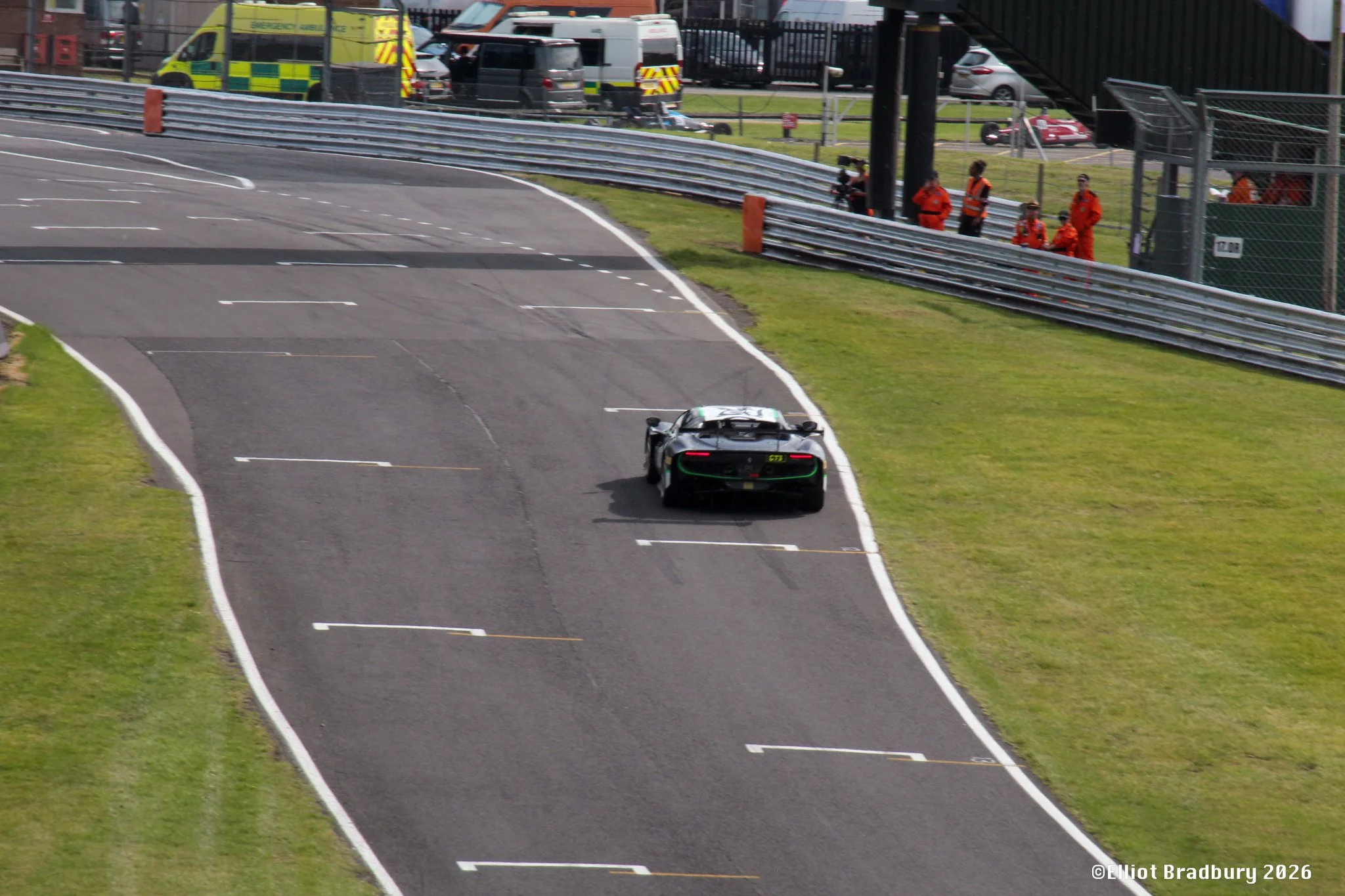 A race track with a sports car driving on it, several emergency medical personnel in orange uniforms are standing and sitting behind a fence, nearby vehicles are parked, and there are other race-related structures and safety barriers visible.