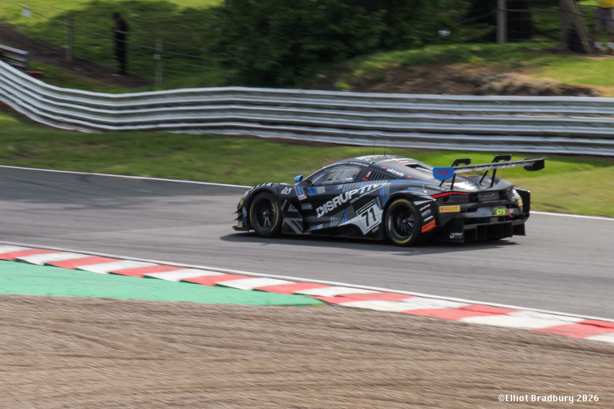 A black race car with blue and white accents speeding on a racetrack with colorful curb markings, surrounded by green trees and a metal guardrail, in what appears to be a motorsport event.