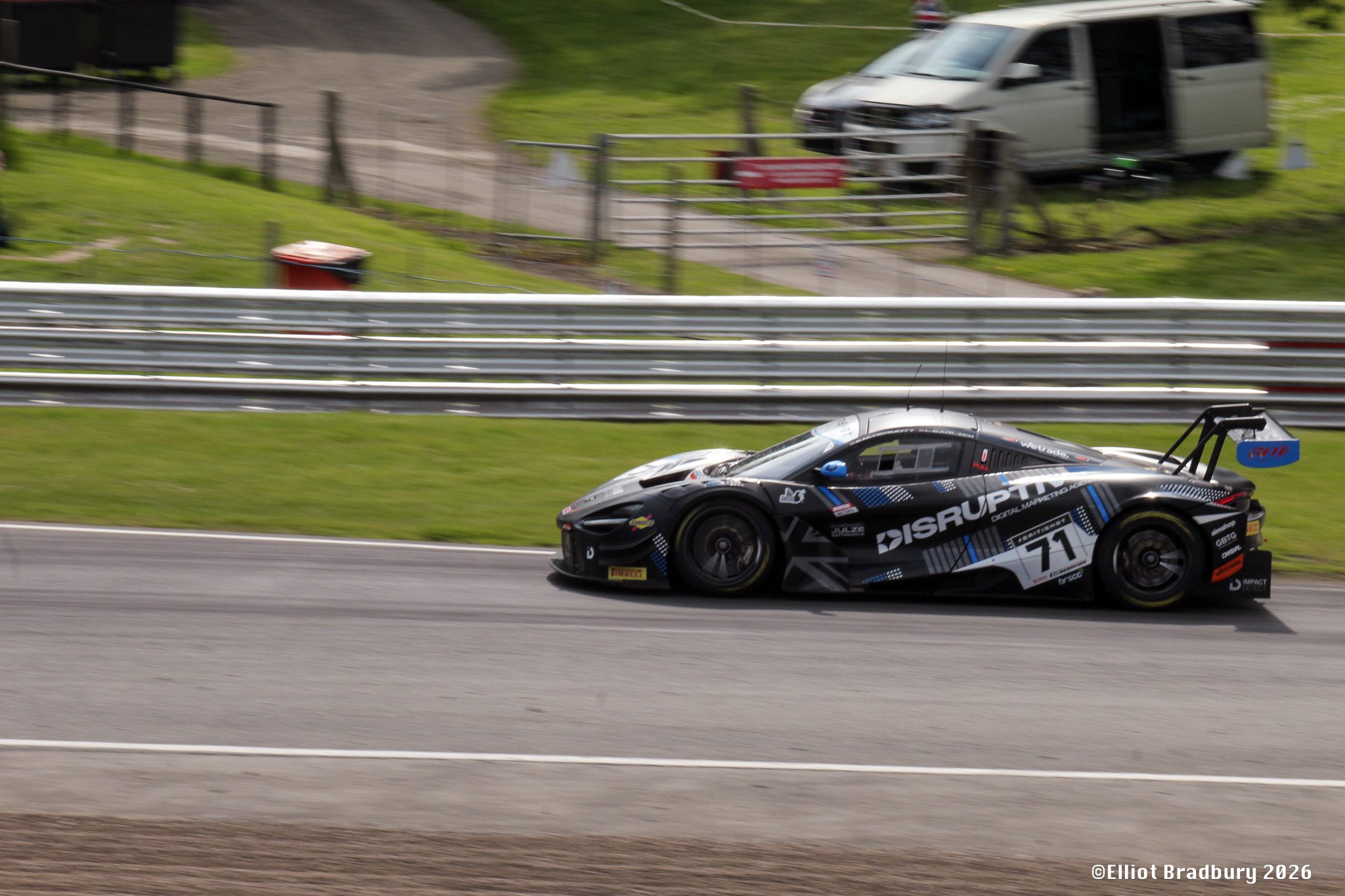 A black race car with blue and white decals speeding on a race track, with blurred background including a van and grassy area.