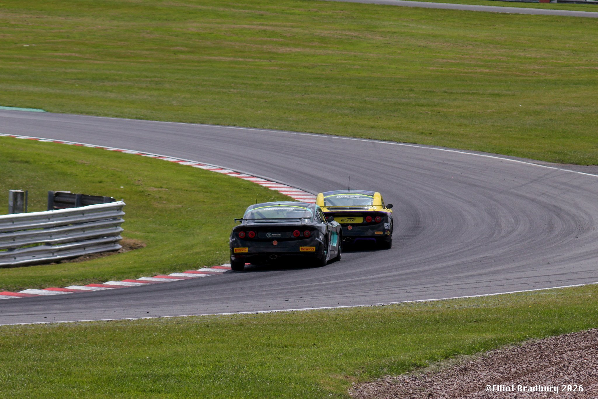 Two race cars navigating a turn on a racetrack surrounded by grass. The cars are close to each other, with one black and the other yellow, both featuring sponsor logos.