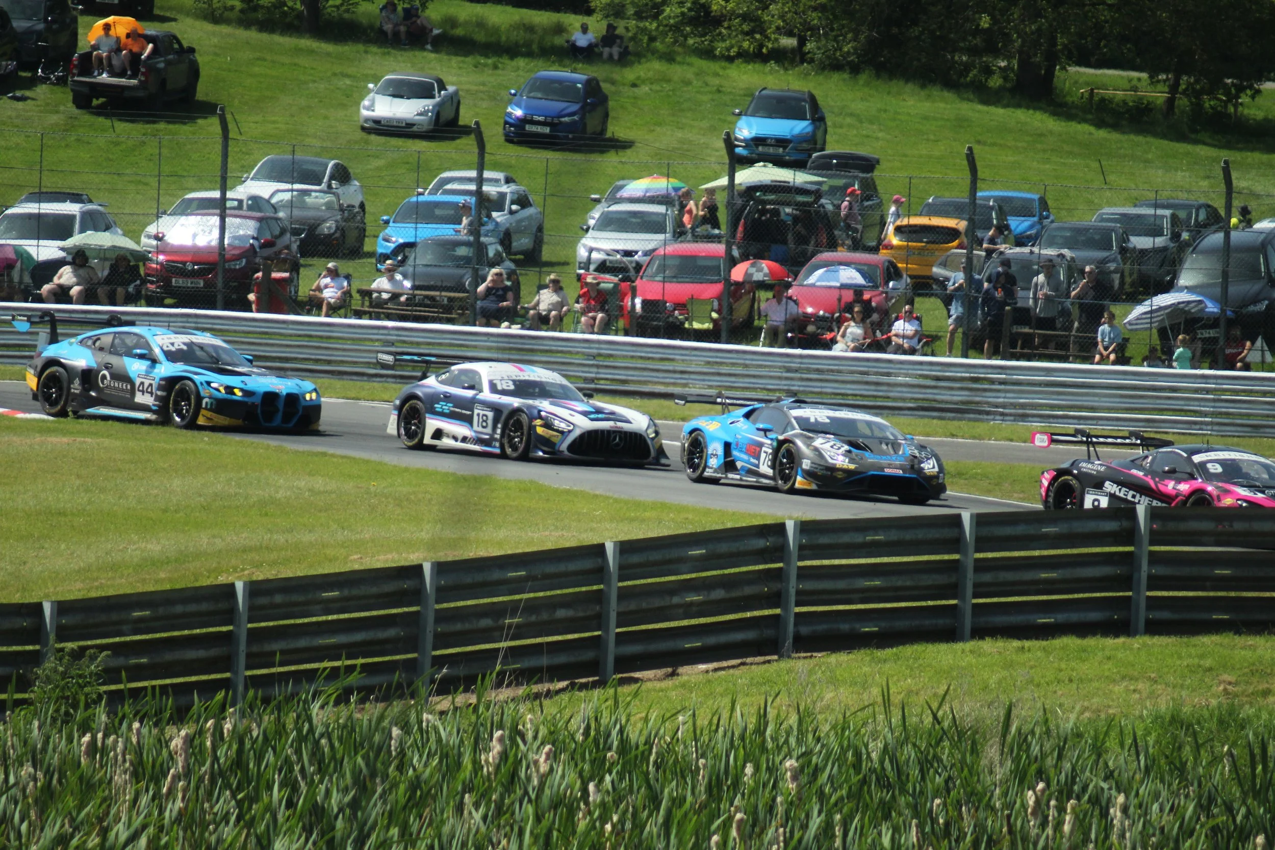 Race cars on a track with spectators sitting on benches and in the grass behind a fence, with cars parked on a grassy hill in the background.