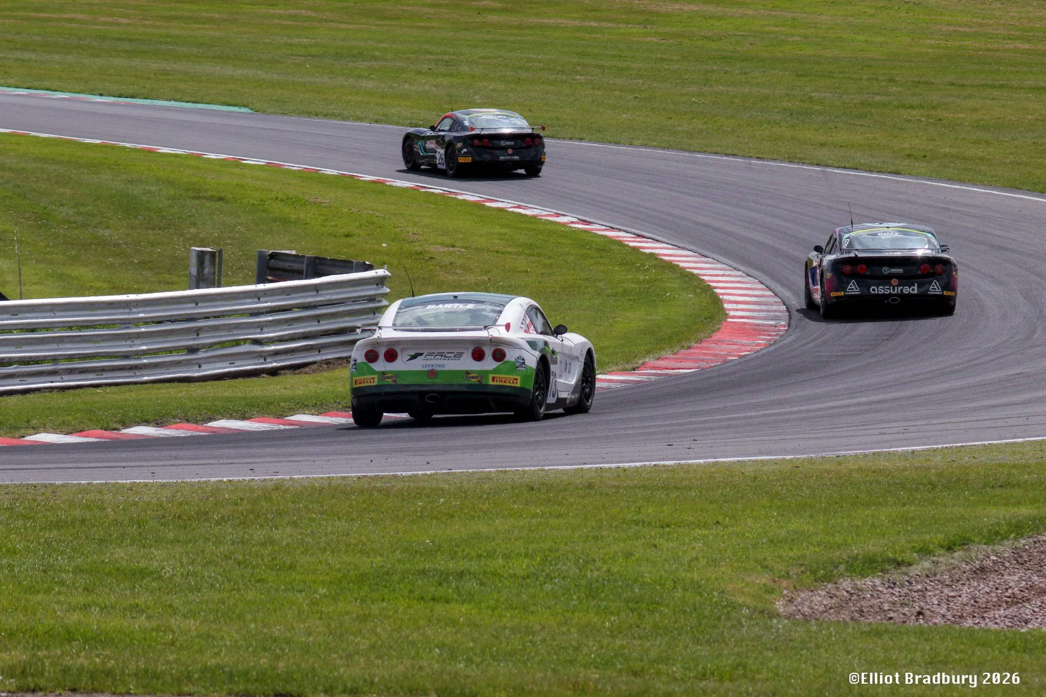 Race cars navigating a turn on a racetrack with red and white curbing, green grass, and guardrails, during daytime.