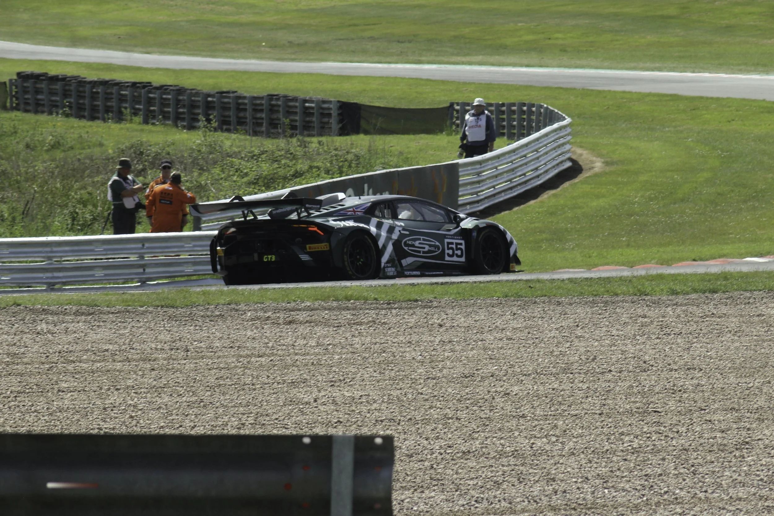 A black race car with the number 55 on its side on a race track, near a safety barrier, with three officials or team members standing nearby watching the car.