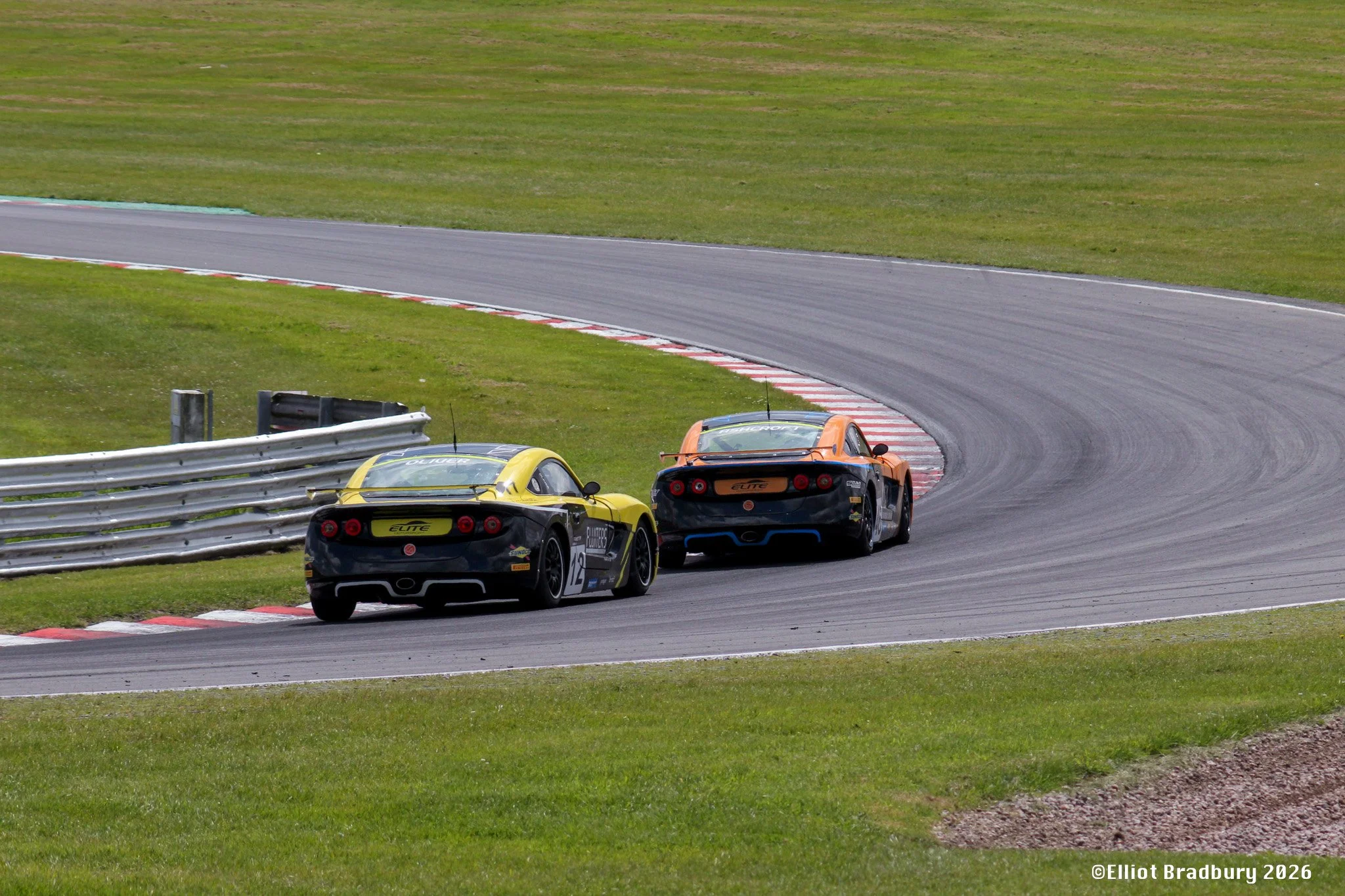 Two race cars in orange and black are navigating a turn on a paved race track, with green grass on both sides and barriers along the edge.