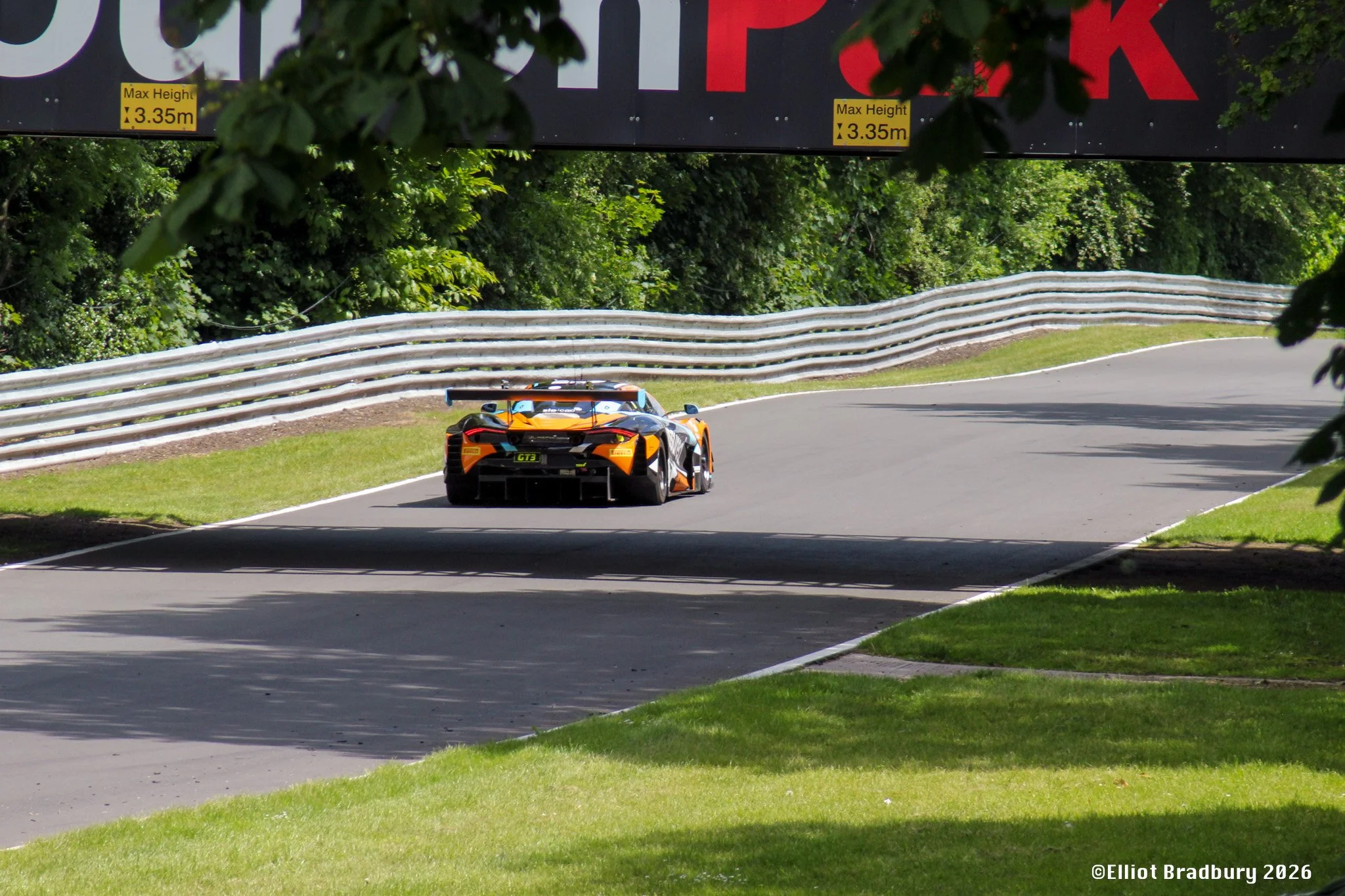 A race car on a winding road surrounded by green trees and grass, with a large overhead sign indicating maximum height clearance of 3.35 meters.