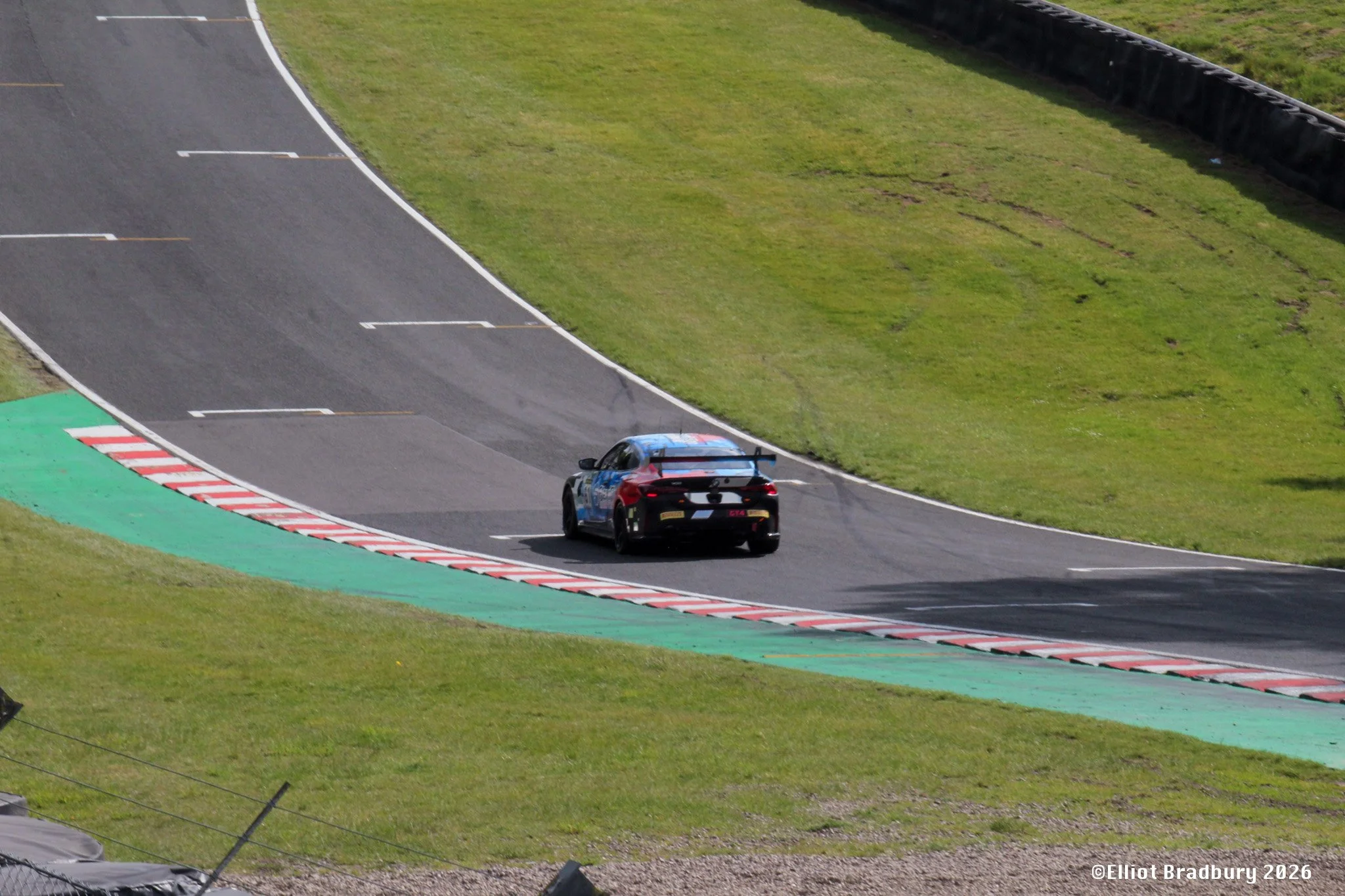 A race car on a curving racetrack with colorful green and red runoff area, surrounded by grassy hills and safety barriers.