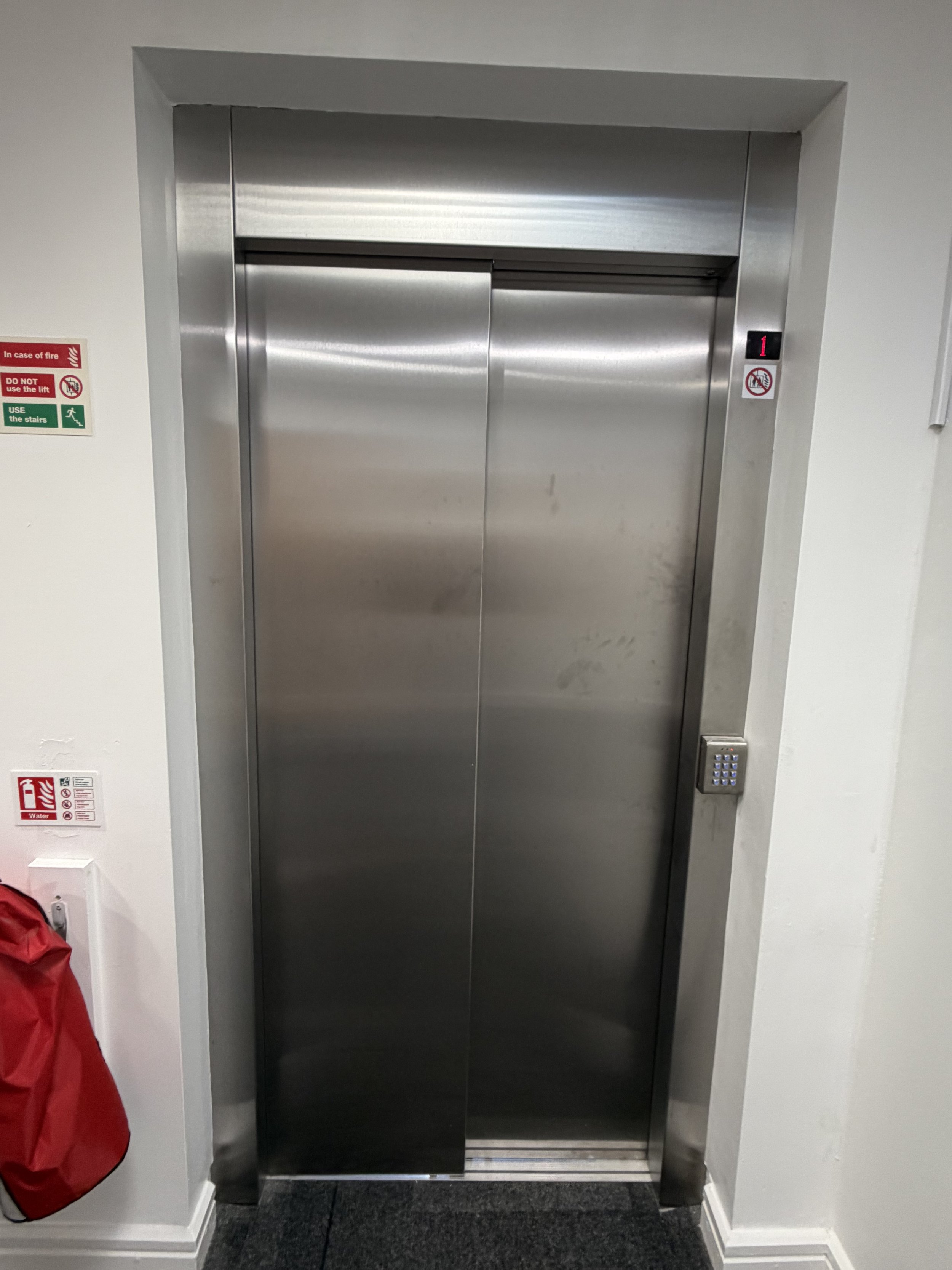 A closed stainless steel elevator door in a white wall with an electronic panel displaying the number 1 on the right side. There are warning signs on the left side of the elevator door, including fire safety and usage instructions.