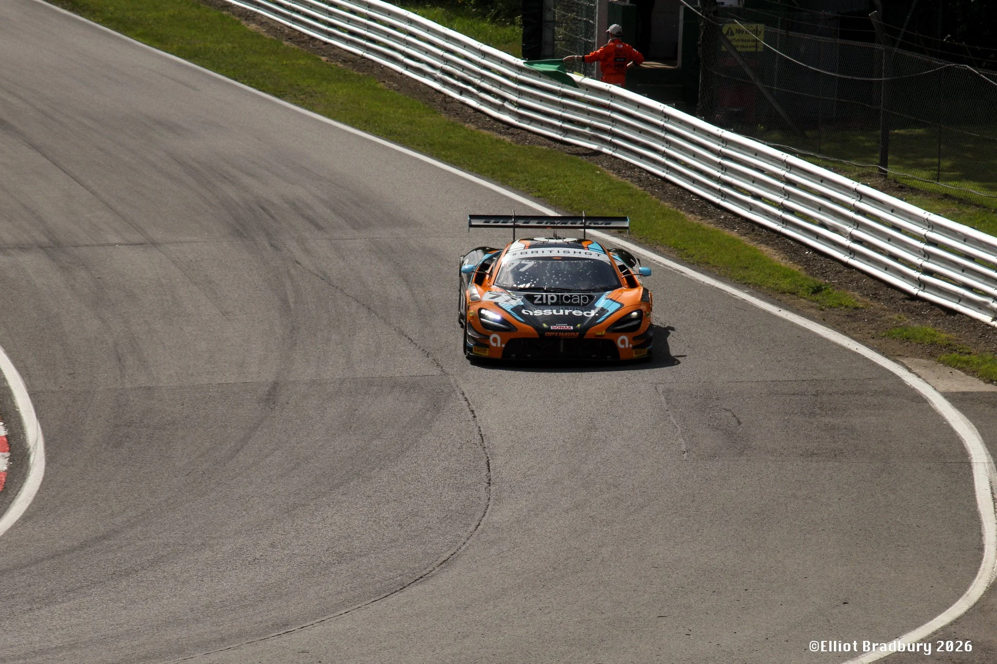 A race car on a racetrack with a person in an orange jacket standing behind the safety fence, holding a green flag. The car has a large rear wing and is painted in orange, black, and blue. The photo is taken from above and to the front side of the ca