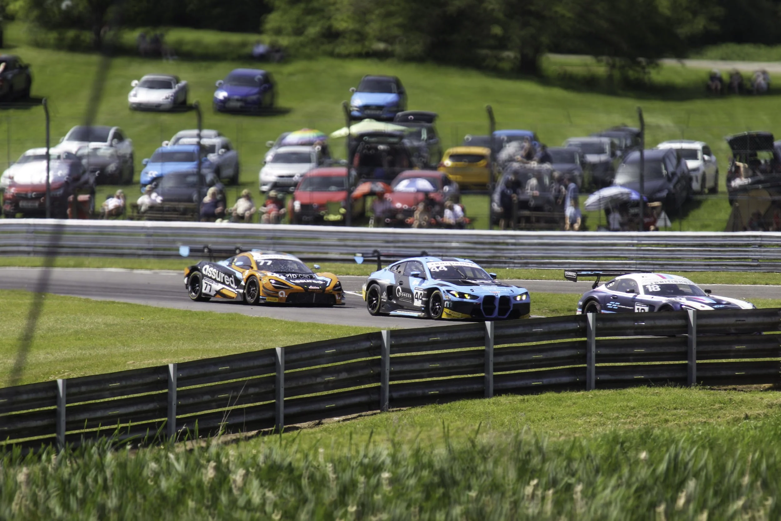 Race cars on a track with spectators in the grass hillside background.