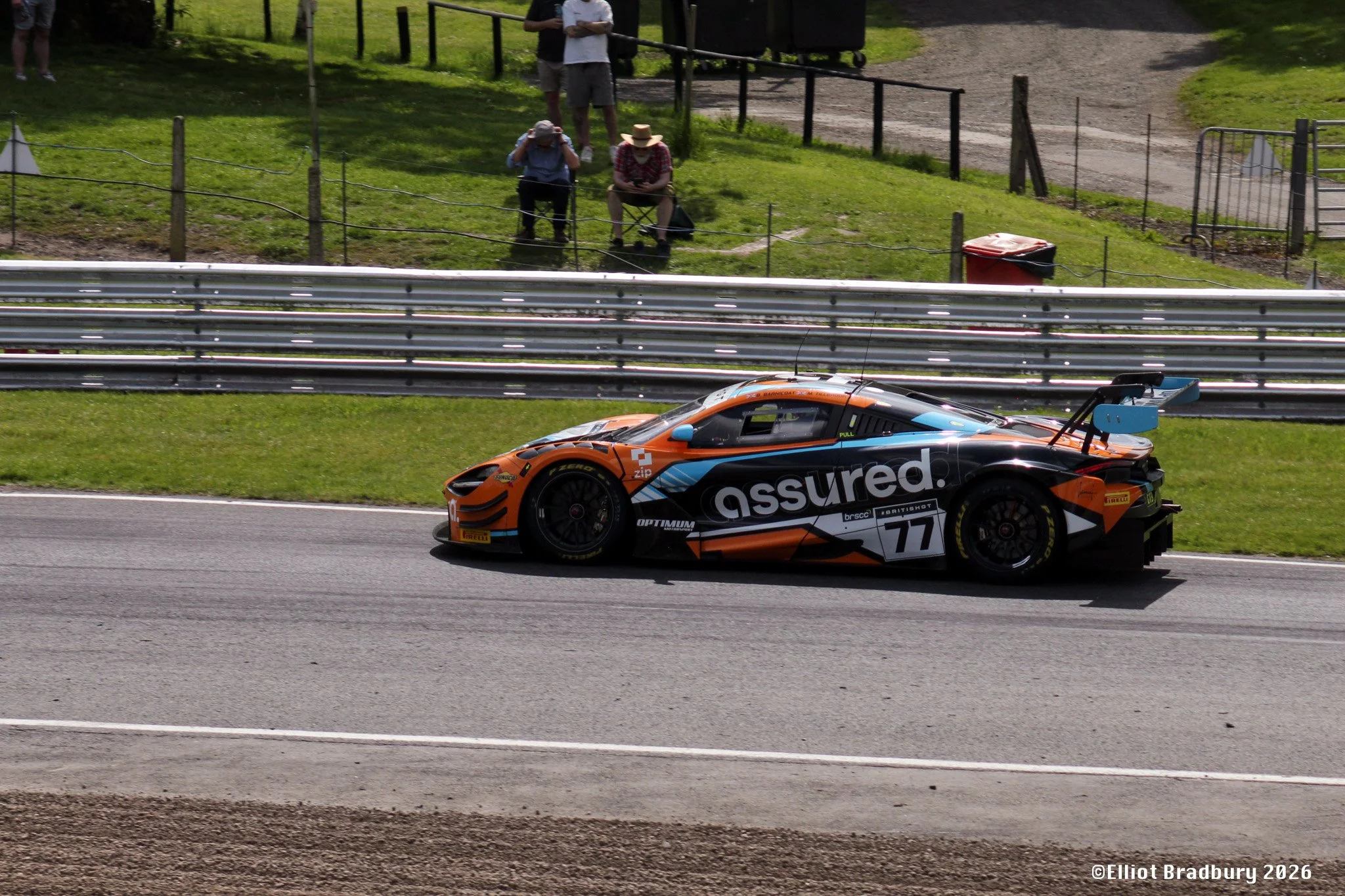 A race car on a track with spectators watching from the grassy area behind a fence.