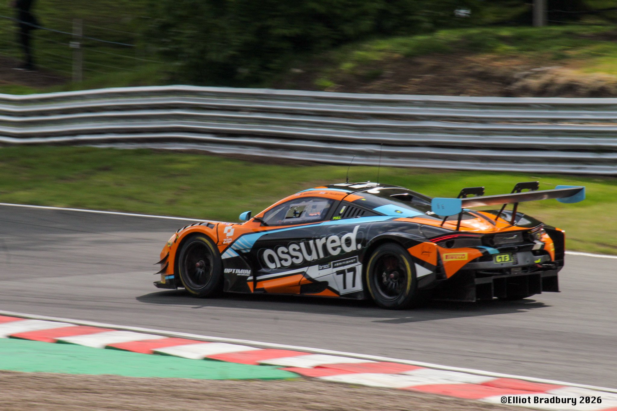 An orange and black race car speeding on a race track, with a large rear wing and sponsor logos, during a motorsport event.