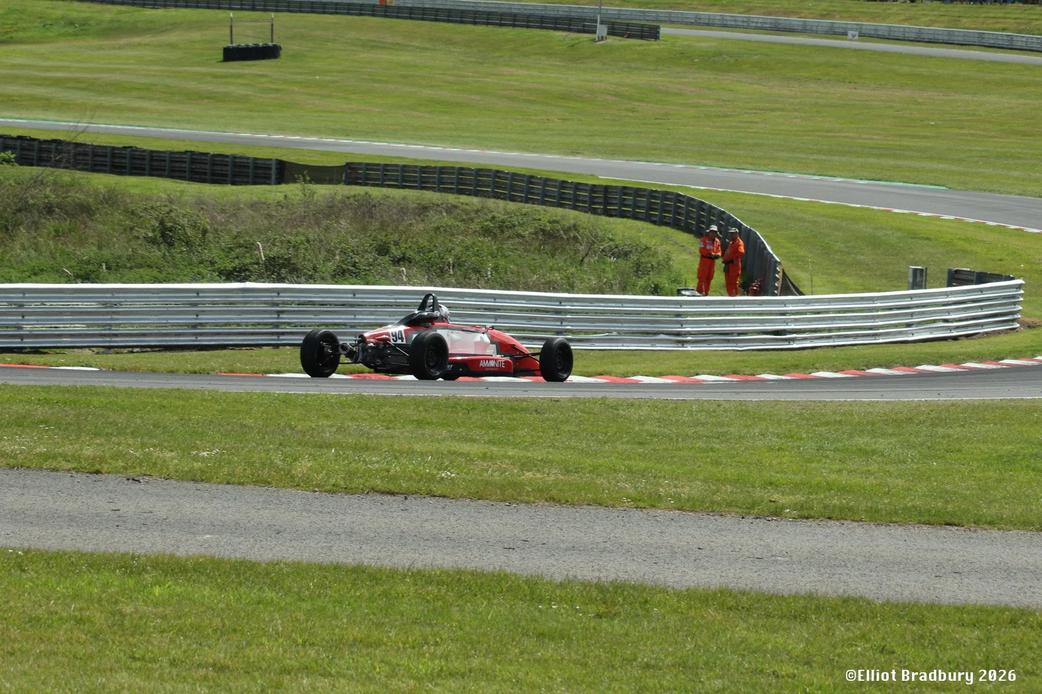 Race car with number 94 on track, two officials in orange uniforms standing near safety barriers, grass and track fencing in background.