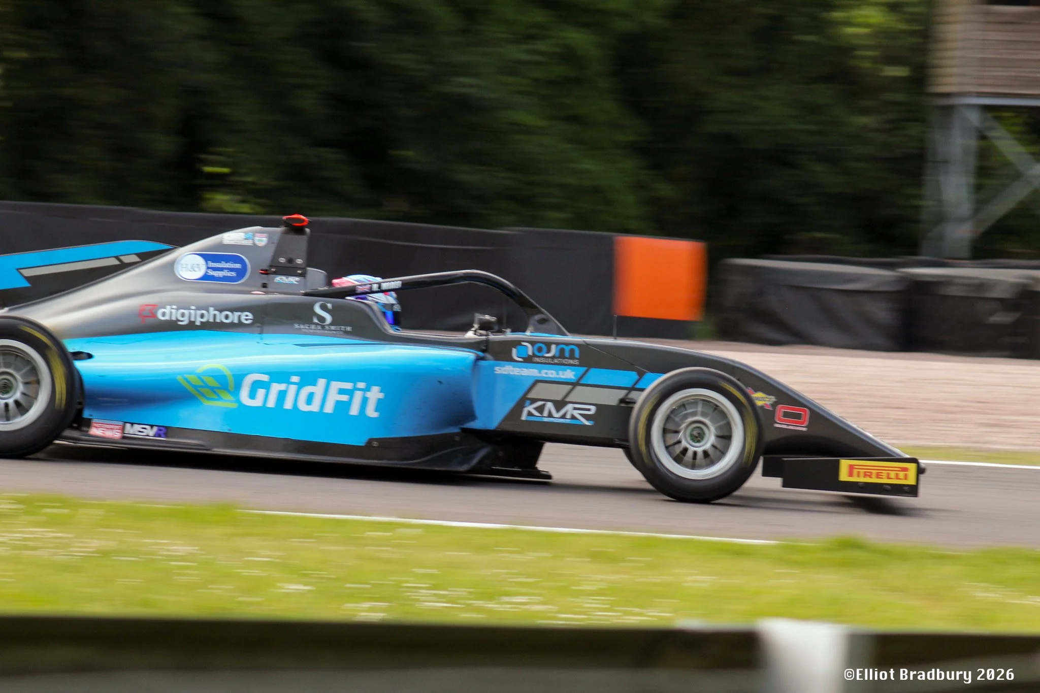 A blue racing car with sponsorship logos, speeding on a race track with greenery in the background.