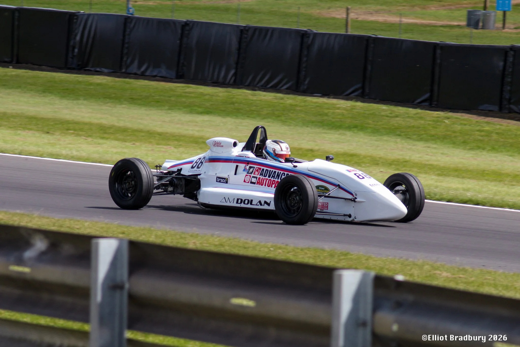 A white racing car with the number 88 on its side and various sponsor logos, including 'Advance Auto Parts' and 'Dolan,' driving on a race track with green grass in the background. Black safety barriers line the track.
