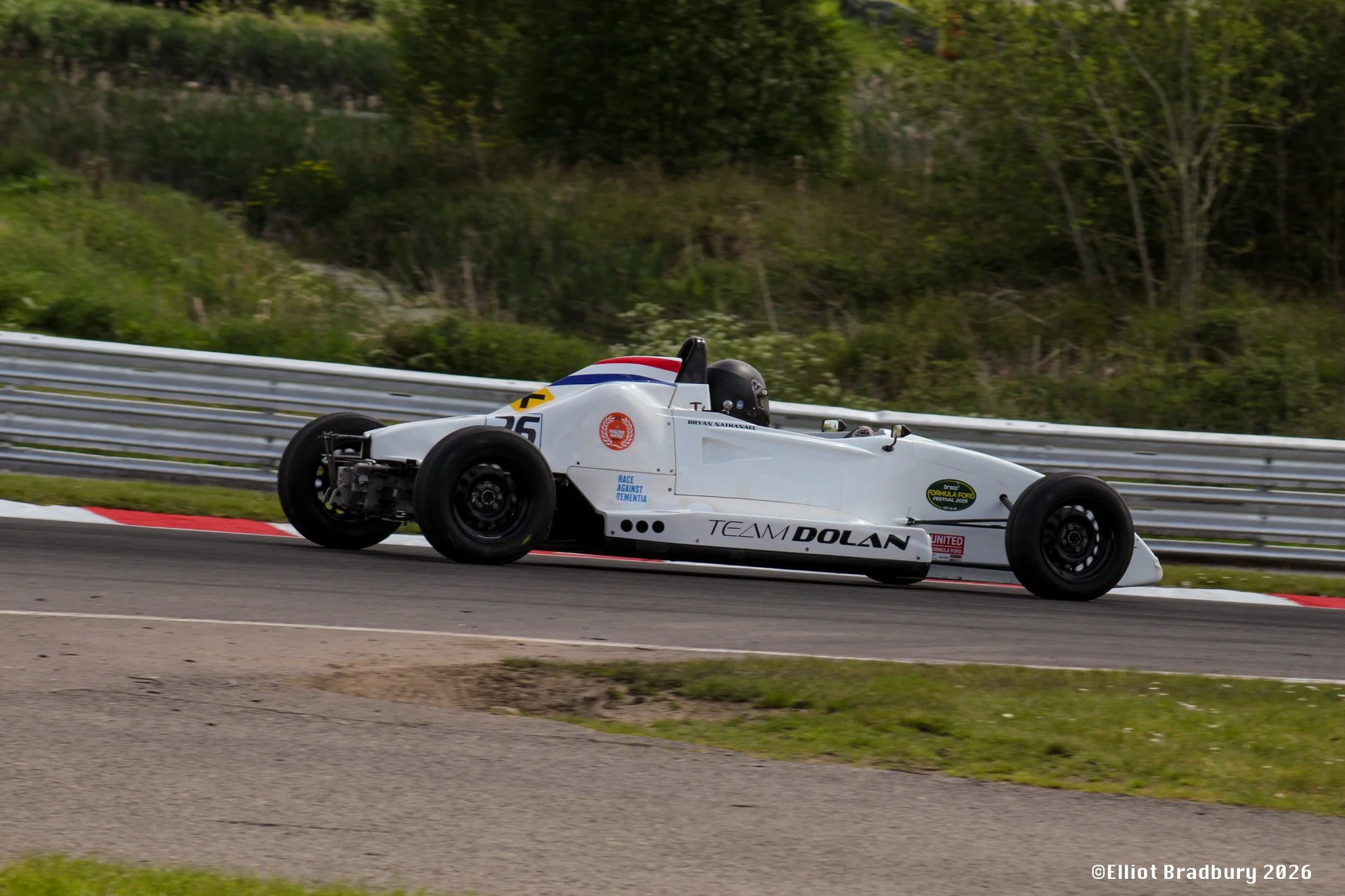 A white racing car with a driver wearing a black helmet on a race track, surrounded by green trees and guardrails.