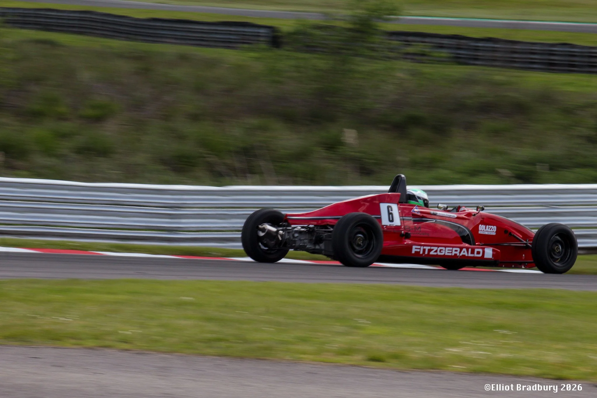 A red open-wheel race car speeding on a racetrack with a blurred background of grass and barriers.
