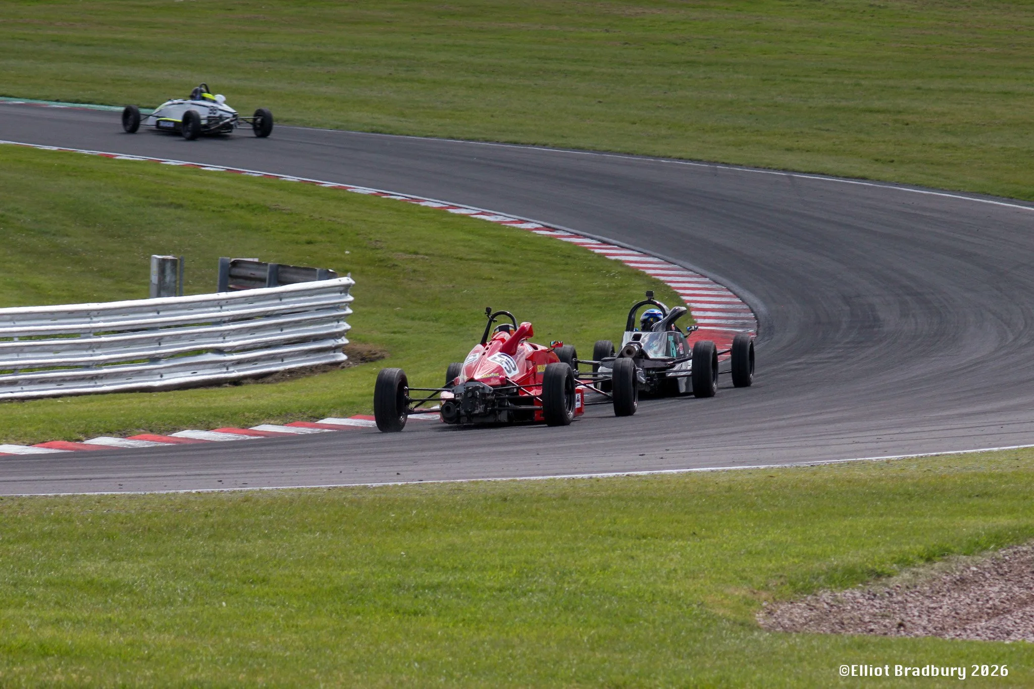 Two open-wheel race cars on a racetrack, with one car in foreground and another ahead around a curve, surrounded by green grass and safety barriers.