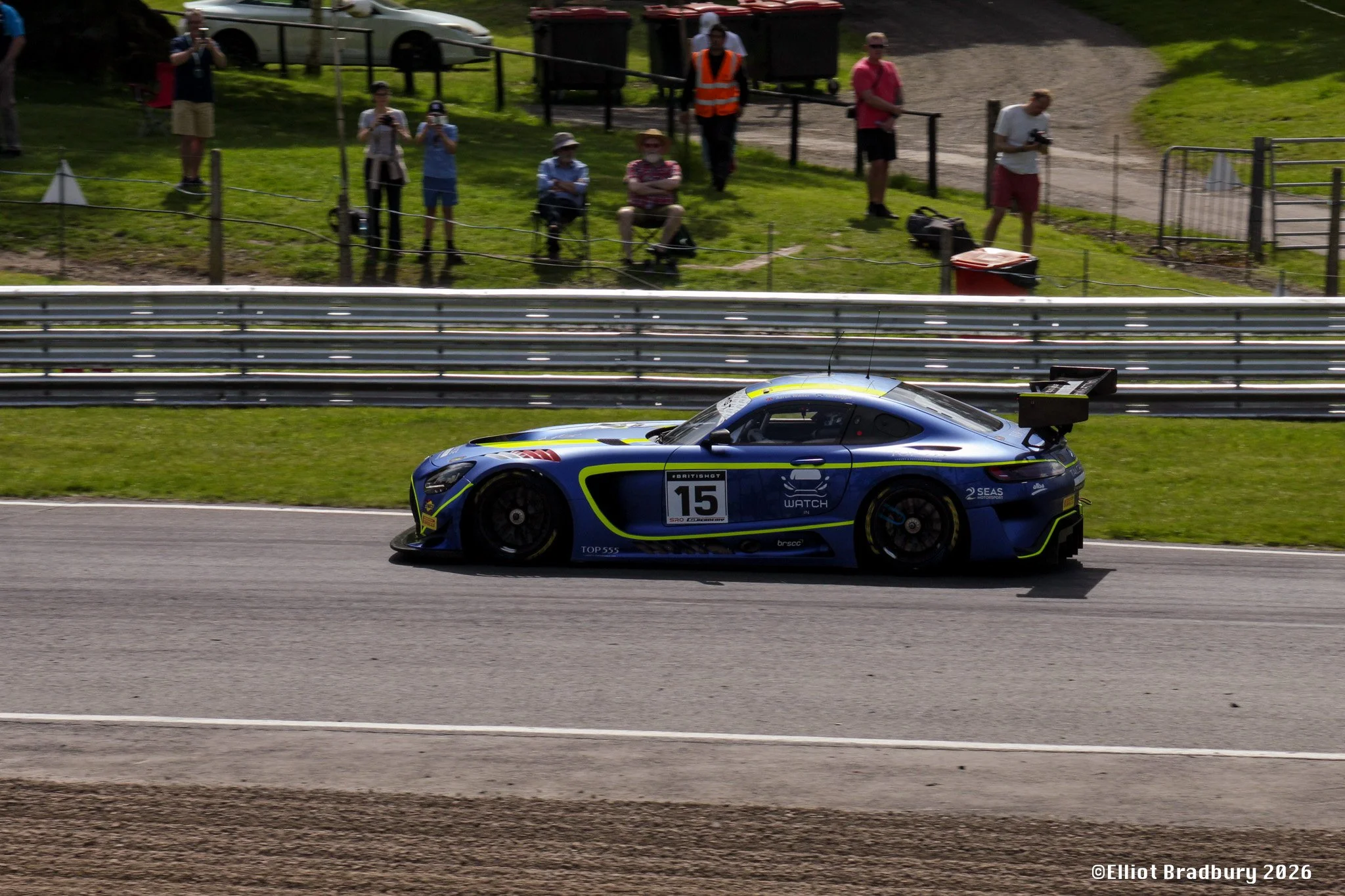 A blue race car with yellow and black accents speeding on a track, with spectators standing behind a fence in the background.