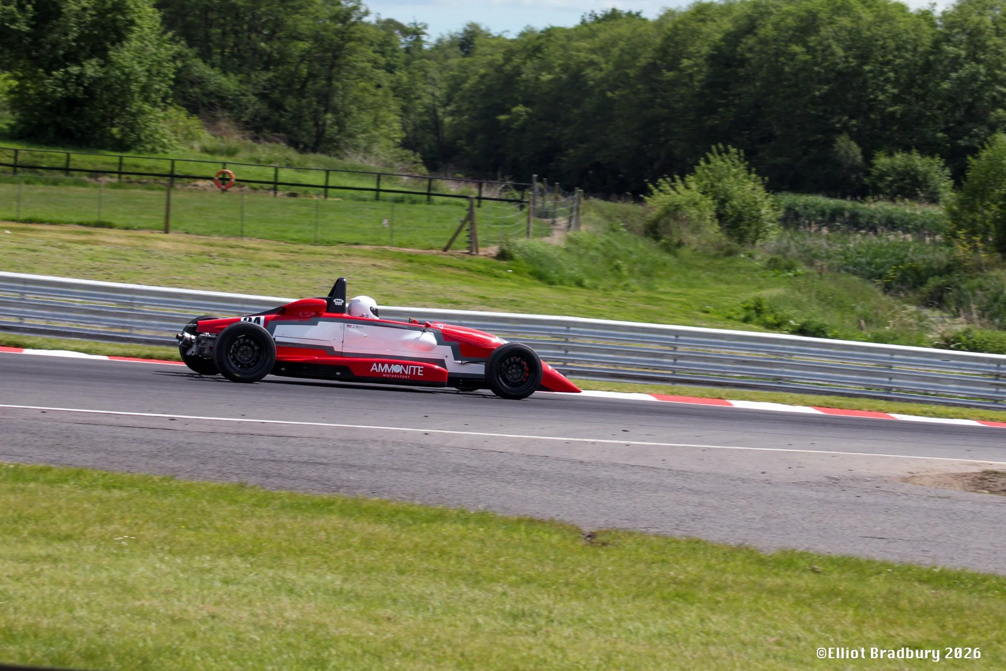 A red and white race car with the number 91 speeds along a curved track surrounded by green grass and trees, with a fence and wooded area in the background.