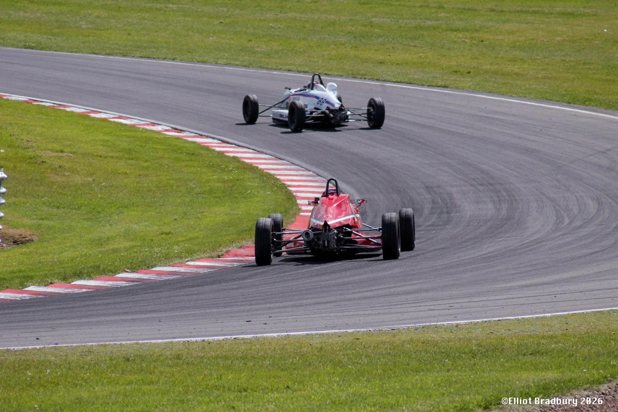 A race car on a racing track, with a red car behind it, both cars are going around a bend on the track with grass on the sides.