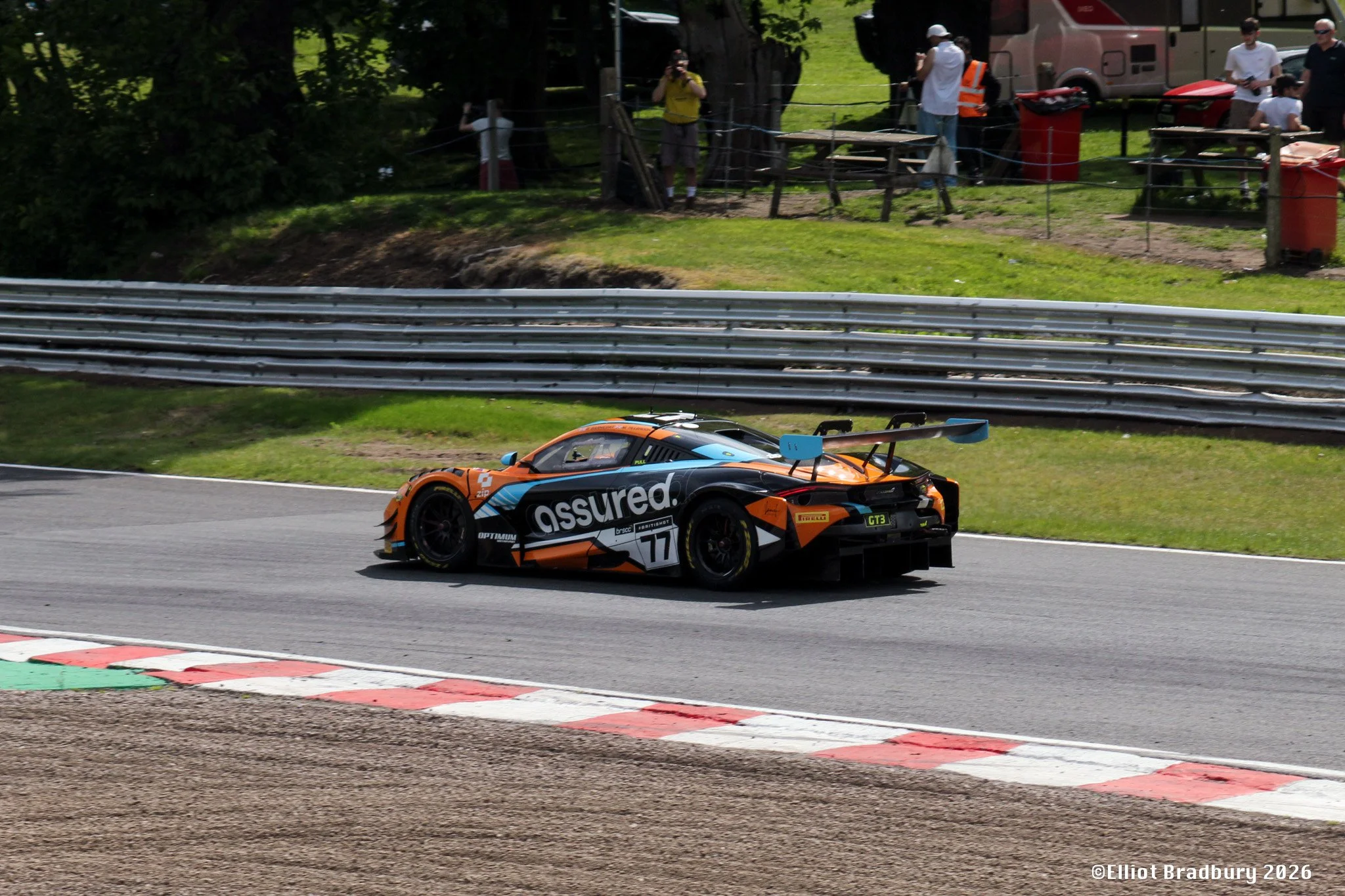 A race car with the number 77 and sponsor logos, moving on a race track with red and white curbing, during a racing event with spectators and staff in the background.