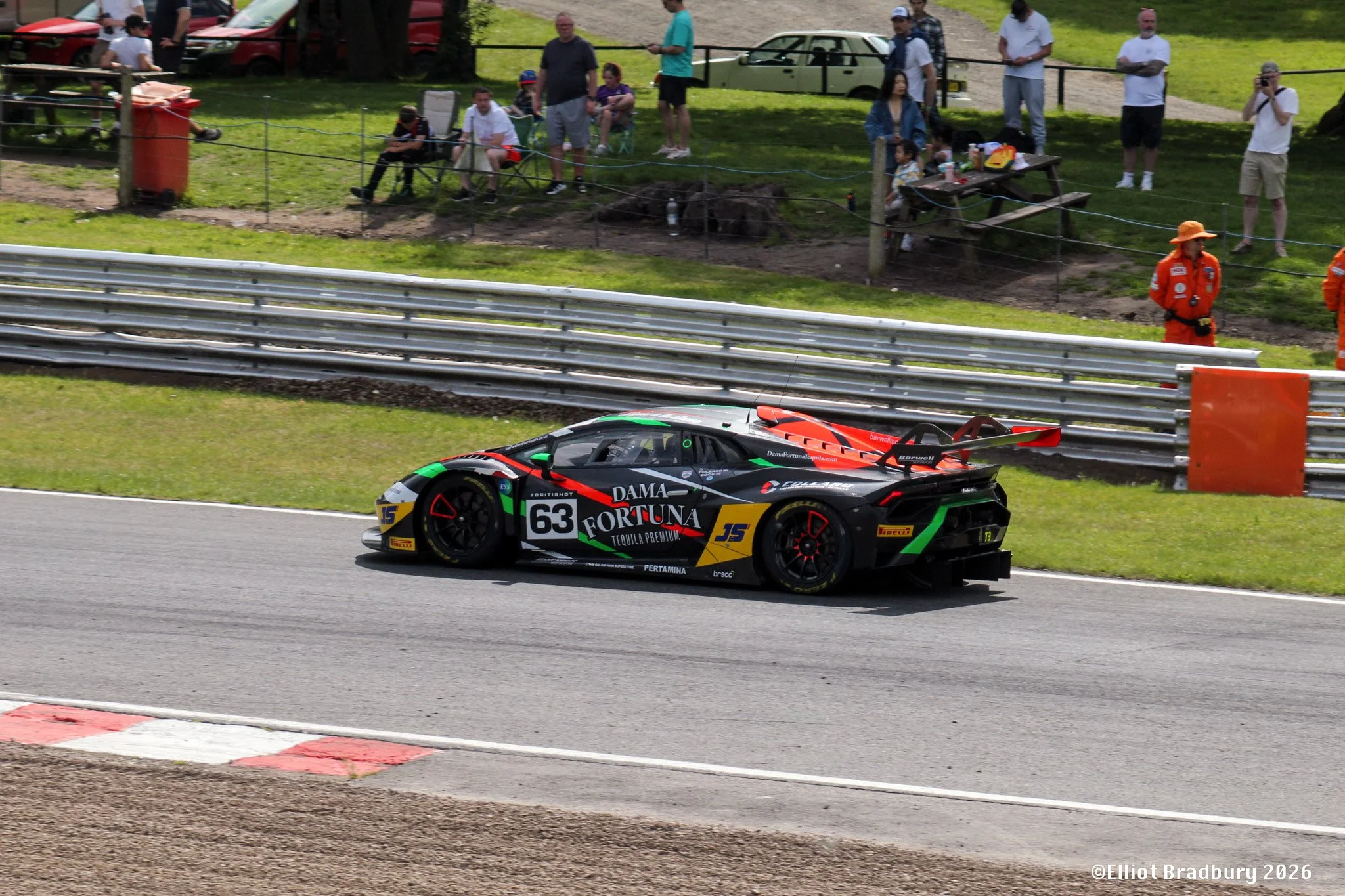 A race car with number 63 on the side, decorated with sponsor logos, speeding along a racetrack, with spectators watching from the grassy hillside behind a barrier and some taking photos.
