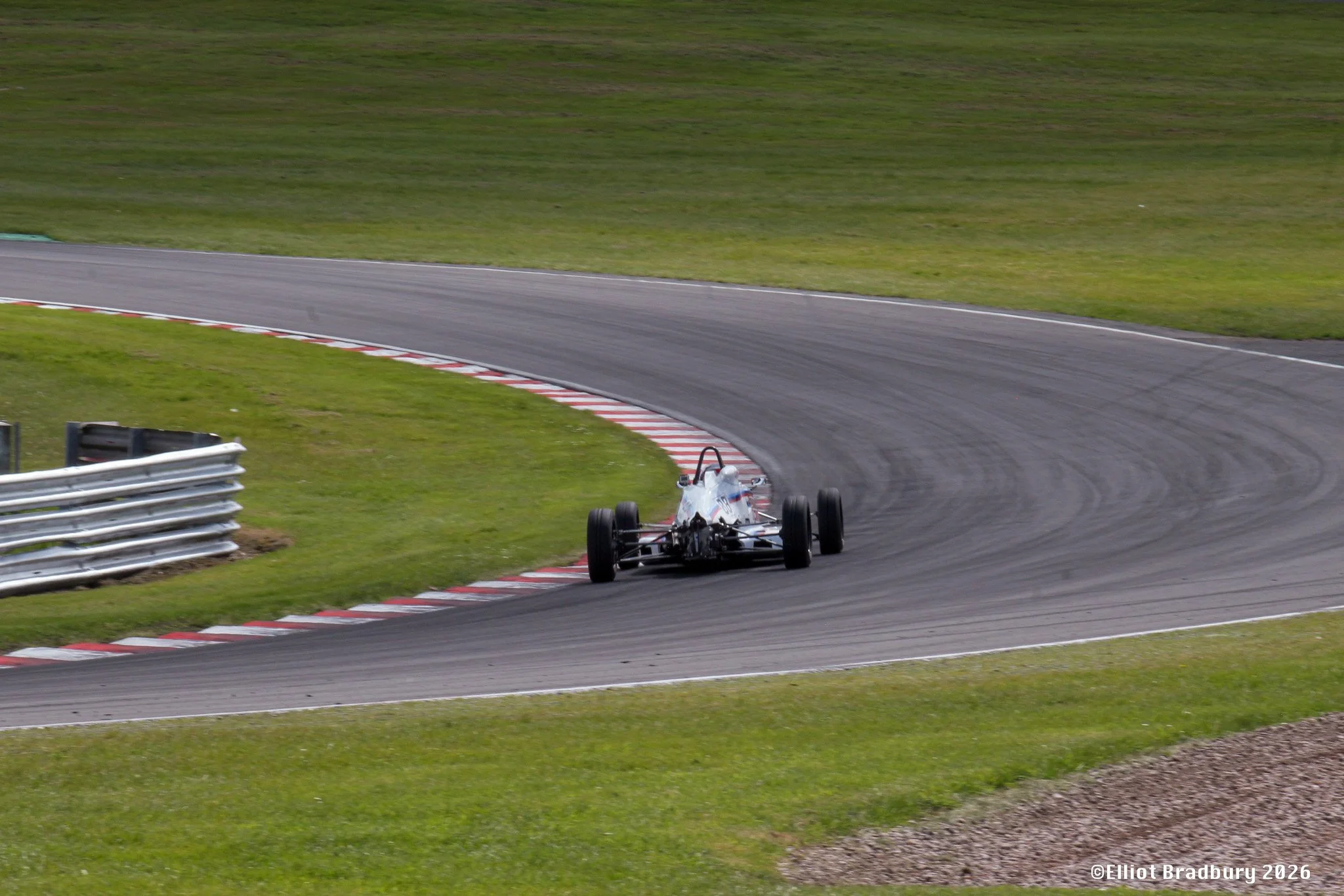 A race car on a racetrack taking a sharp turn with grass on either side and safety barriers at the edge of the track.