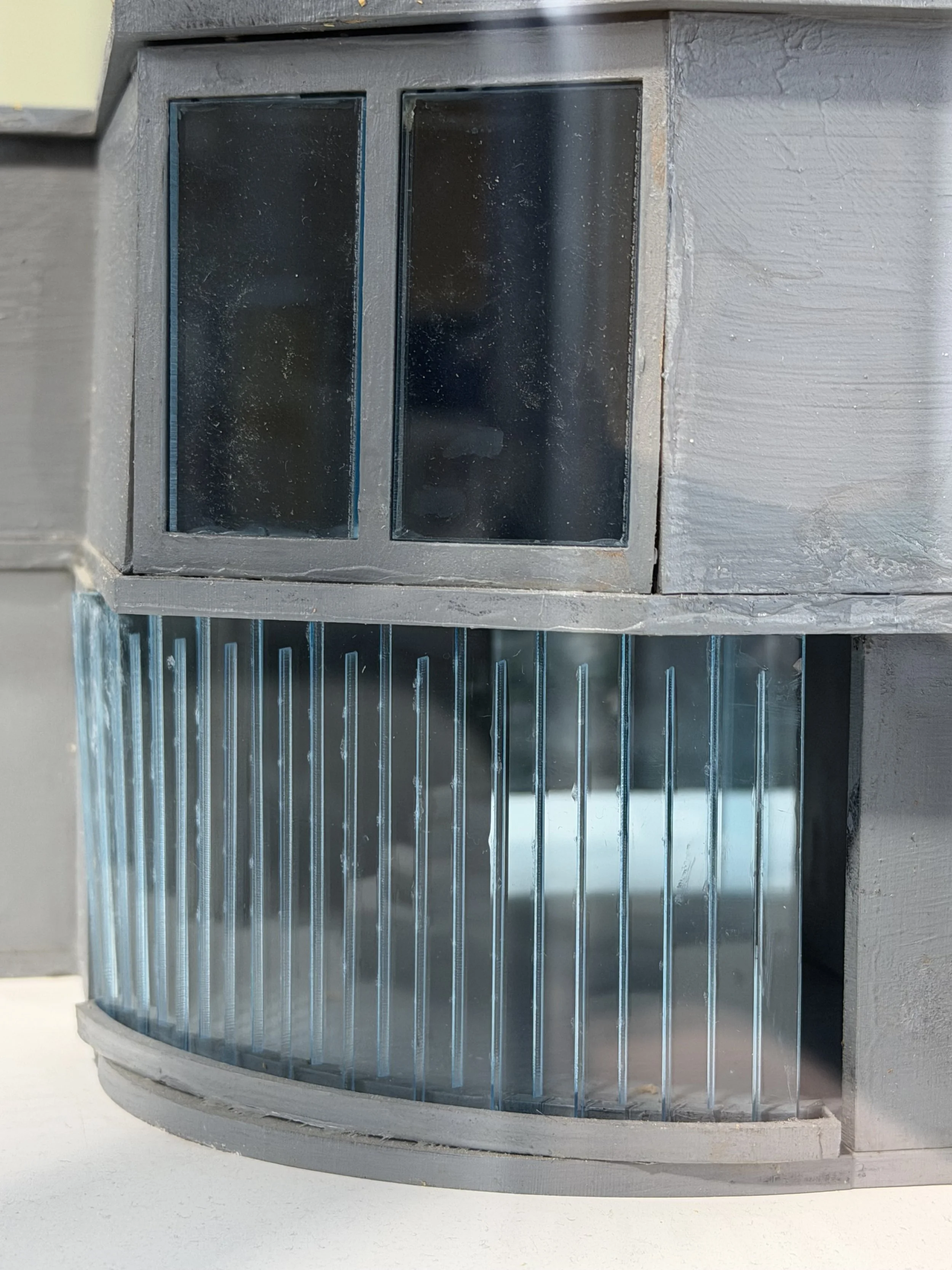 A curved gray metal cage vent with blueish metal bars and a dark background, situated on a gray wall.