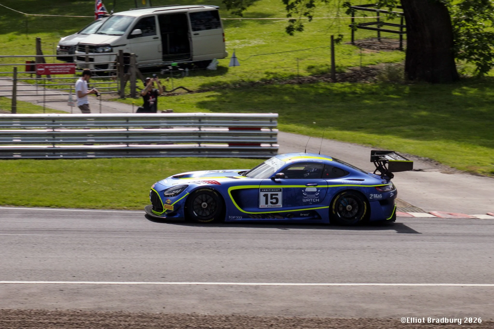 A blue race car number 15 speeds along a track, with a white van and two people in the background near a wooded area.