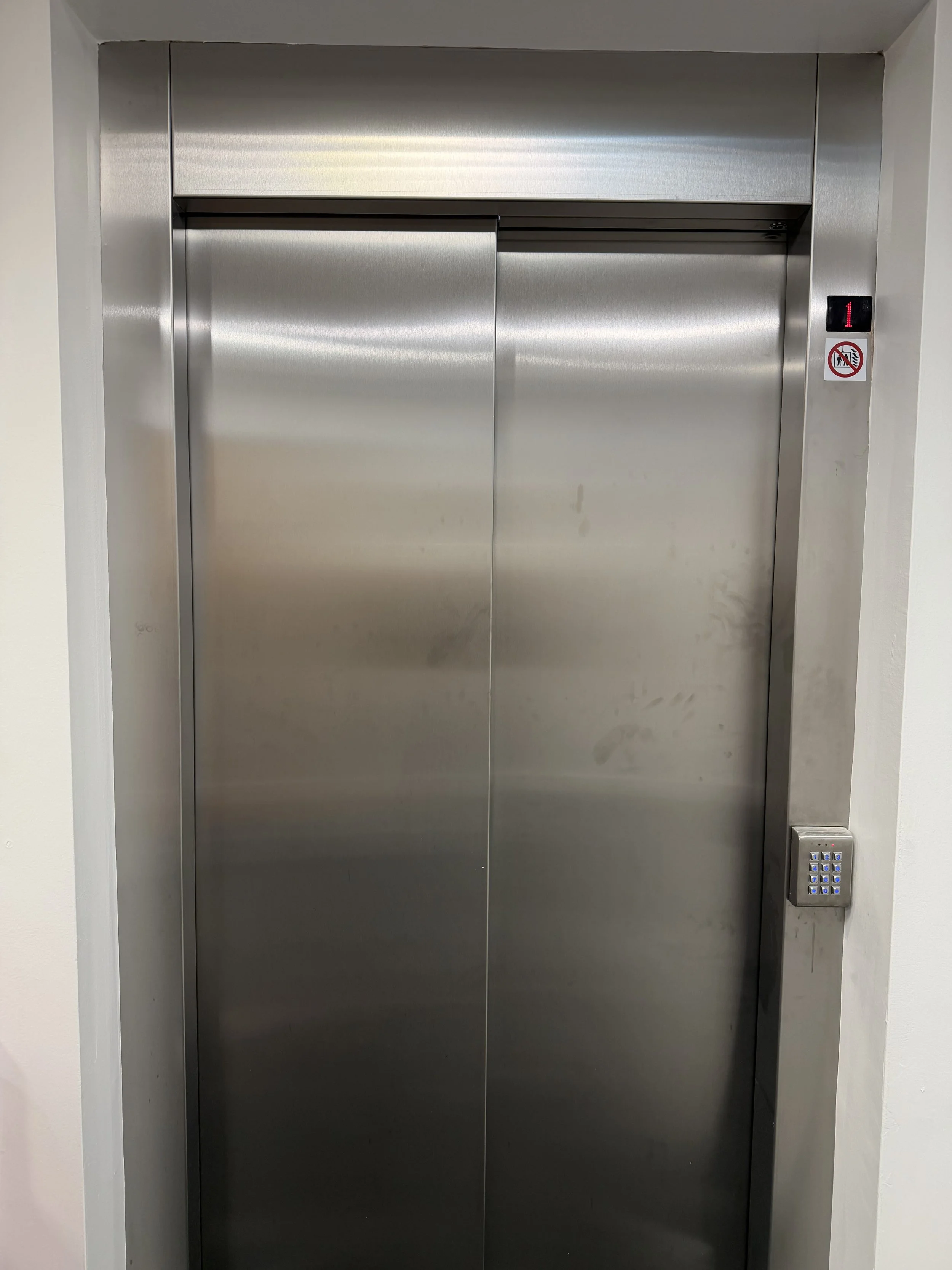 Closed elevator doors made of stainless steel with a keypad to the right and no smoking sign below a digital floor indicator showing level 1.