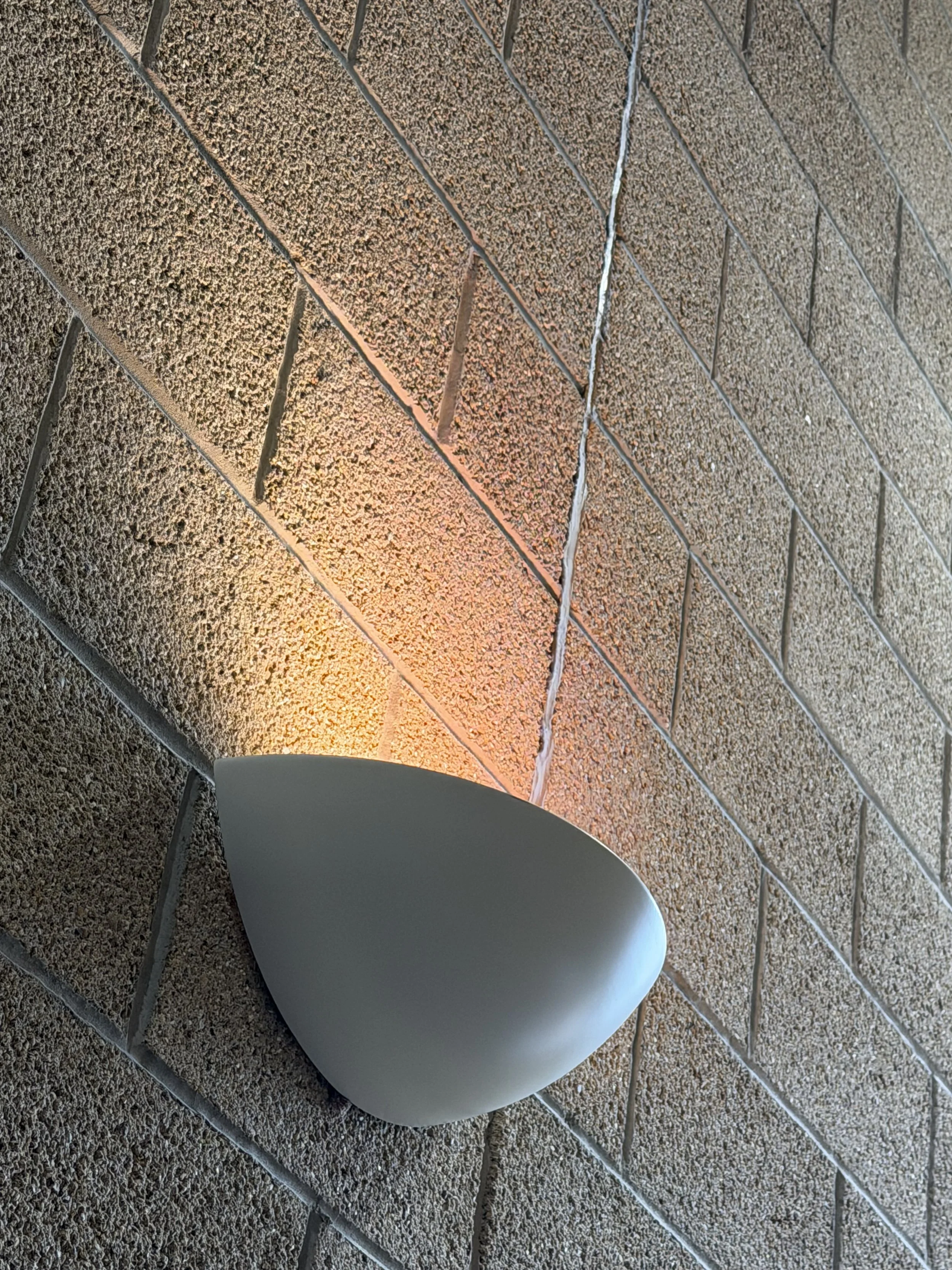 A white, modern, curved lamp with warm lighting, placed against a textured brick wall.
